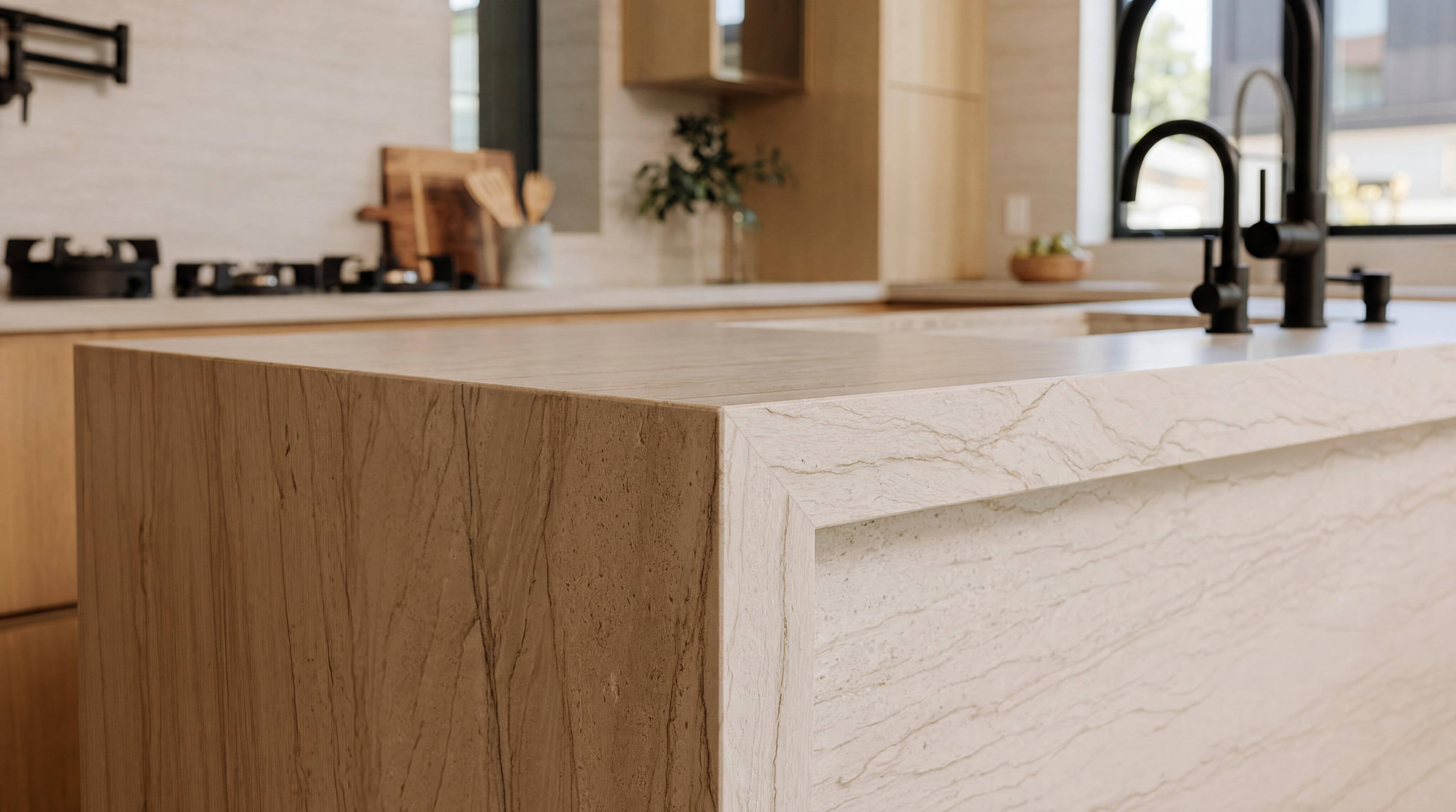 This image showcases a sophisticated kitchen island featuring a waterfall edge with natural, textured stone veining, which provides a high-end, organic aesthetic. In the background, modern matte black fixtures and clean-lined cabinetry create a contemporary, minimalist design. The warm, neutral color palette integrated with premium materials highlights the luxury craftsmanship of the space.