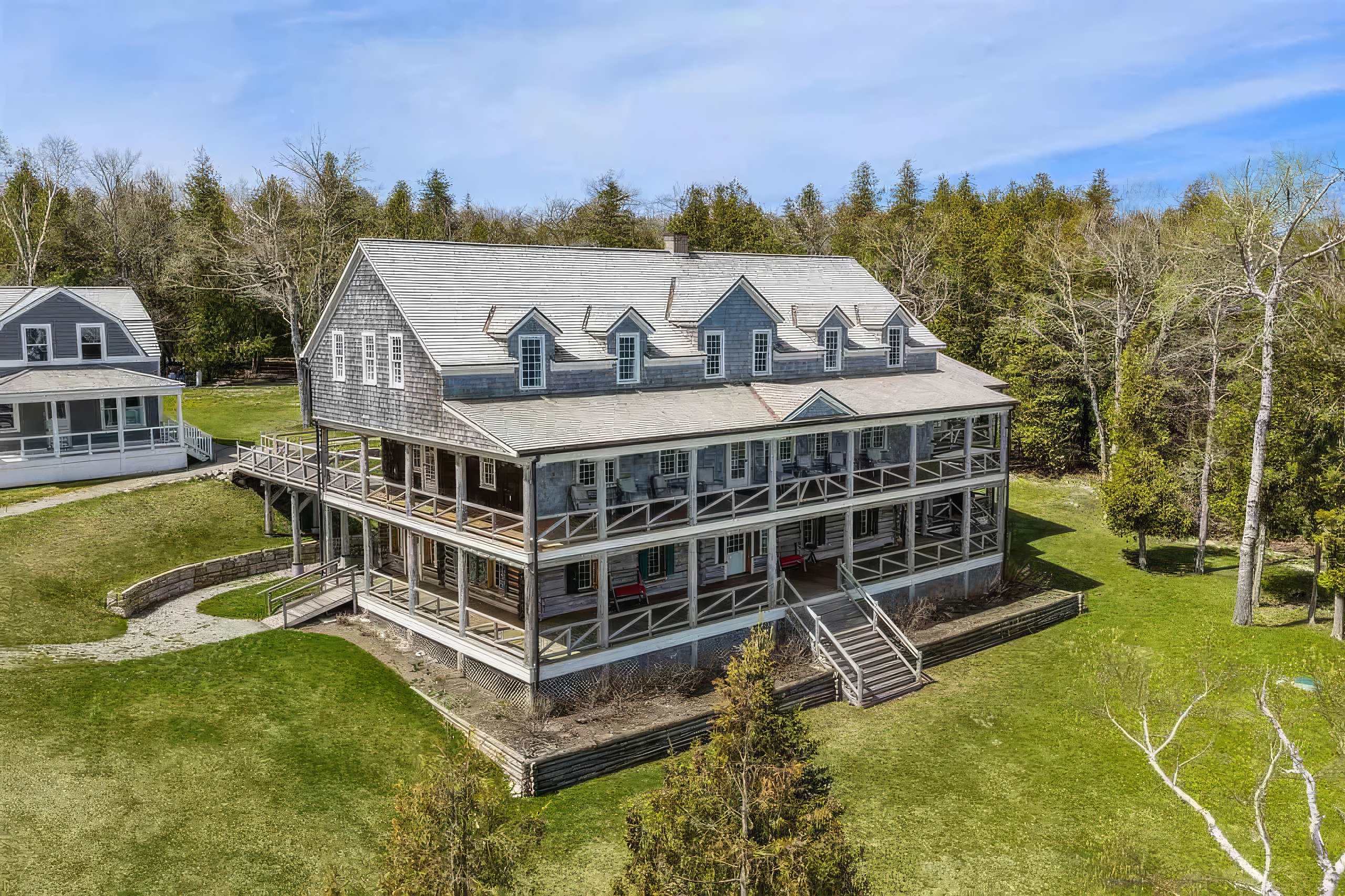 This aerial view captures a large, multi-story shingled home featuring expansive wrap-around porches on two levels. The structure is set against a backdrop of dense forest, with a well-maintained lawn and a stone retaining wall defining the landscape. The perspective highlights the home's classic architectural design, including multiple dormer windows and a symmetrical facade.
