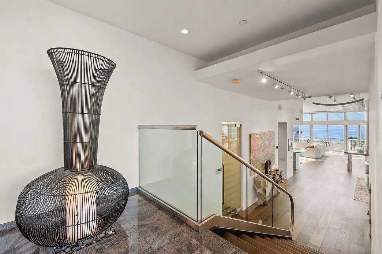 This interior view captures a sophisticated entryway and staircase area featuring a striking, oversized black wire-frame sculpture that houses a glowing light fixture. A glass-paneled railing lines the staircase, which leads down to a bright, open-concept living space with light wood flooring and expansive windows overlooking the ocean. The aesthetic is modern and minimalist, emphasizing clean lines, high ceilings, and a seamless transition between the elevated landing and the main living area.
