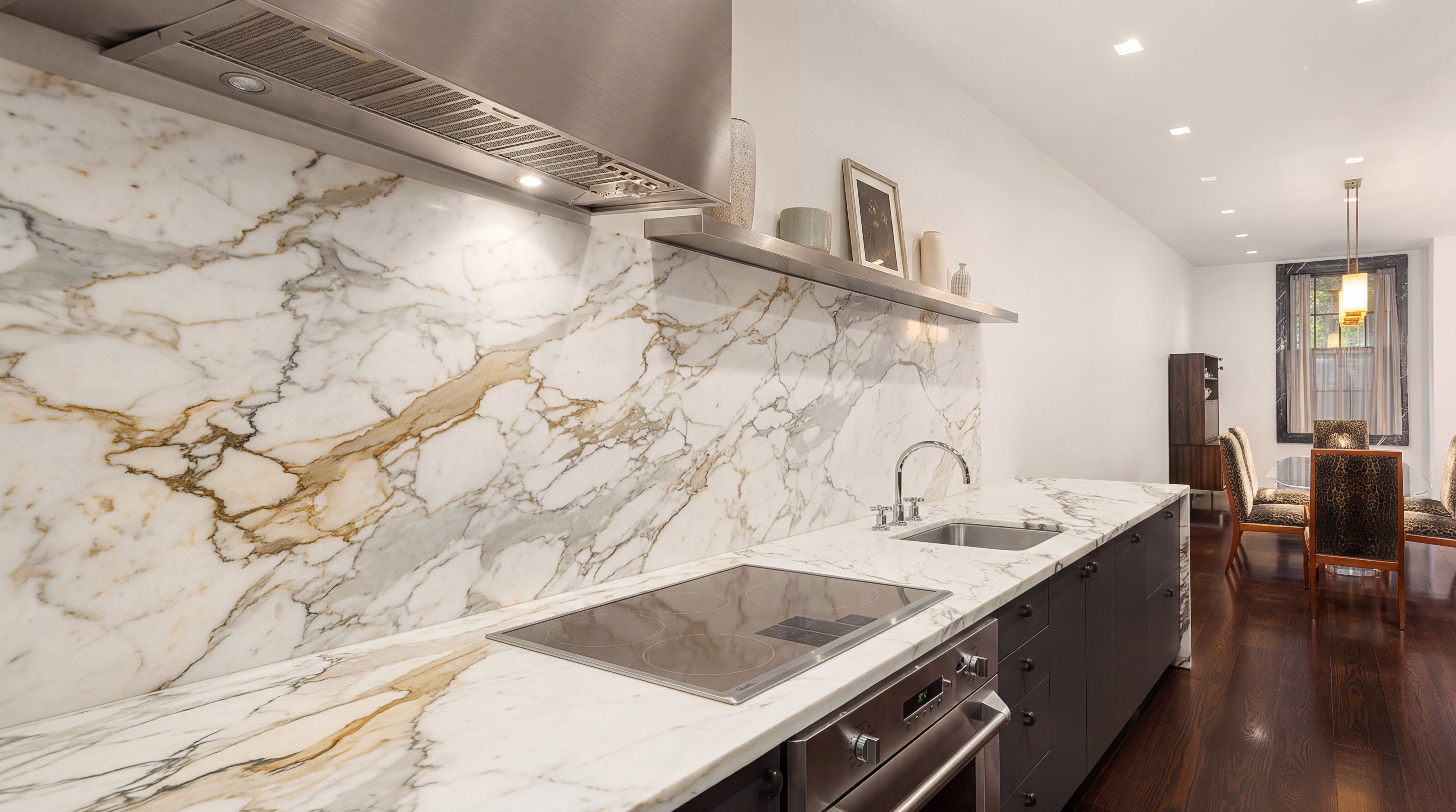 This modern kitchen features luxurious white marble countertops and a matching full-height backsplash, providing a sophisticated, high-end aesthetic. It is equipped with a professional-grade stainless steel vent hood, an electric cooktop, and contrasting dark cabinetry, all beautifully illuminated by sleek task lighting. The seamless layout opens toward a dining area, emphasizing a spacious, elegant design suitable for refined living.