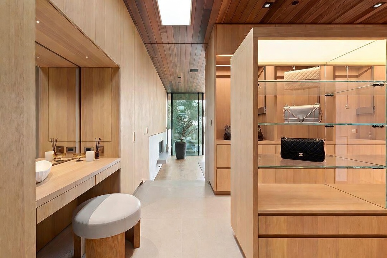 This sophisticated walk-in closet features warm, light-toned wood cabinetry and a minimalist aesthetic. A dedicated vanity area with a mirror and stool sits opposite a glass-enclosed display case showcasing luxury handbags, while a skylight and recessed lighting create a bright, airy atmosphere. The perspective looks down a clean, modern corridor that leads toward a glass-walled view of the outdoors.