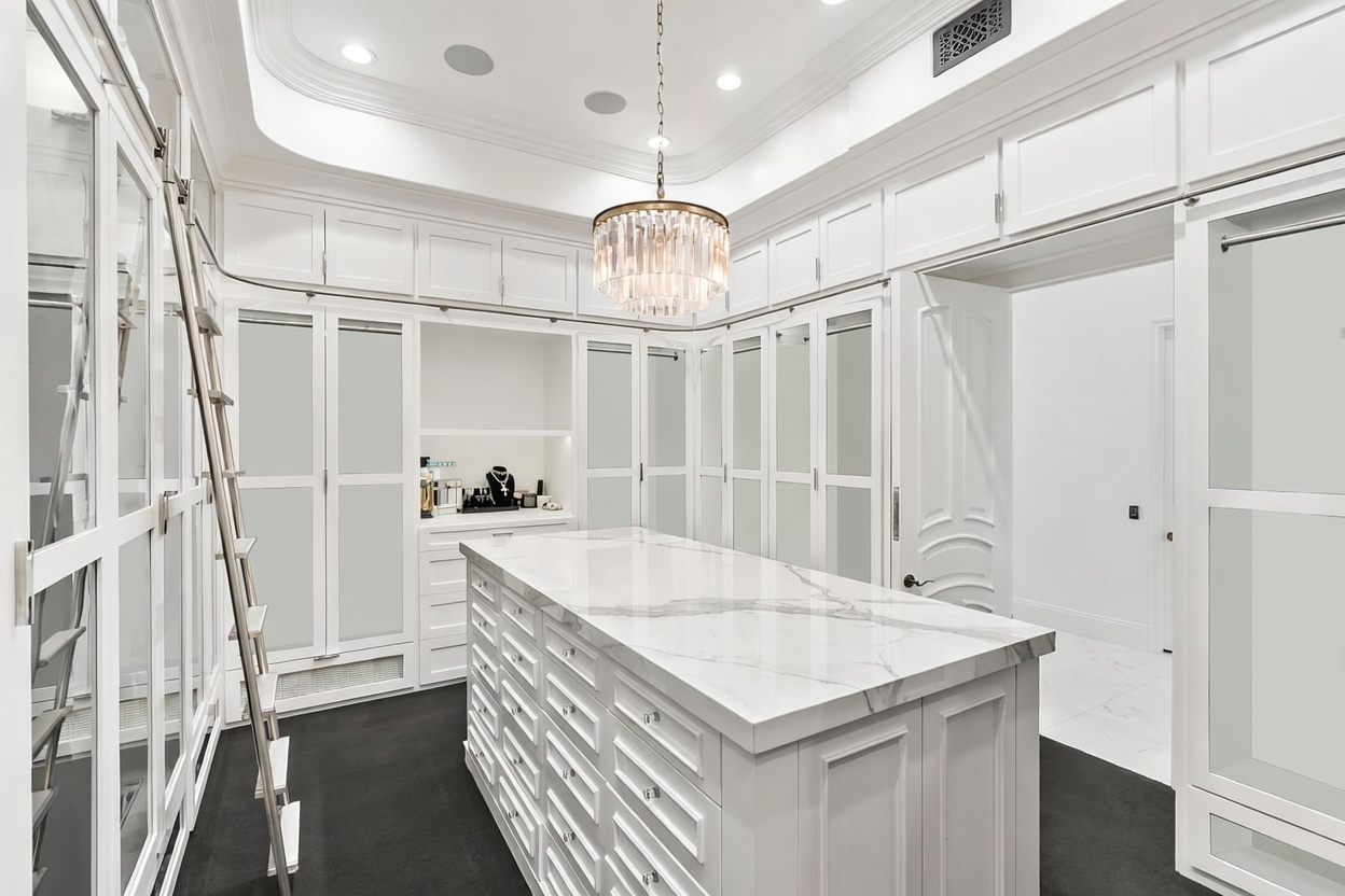 This is a luxurious walk-in closet featuring custom white cabinetry with glass-fronted doors and ample storage space. A central island with a marble countertop provides additional surface area, while a crystal chandelier adds a touch of elegance. A rolling ladder provides access to the upper cabinets, and a television is mounted above a display area.