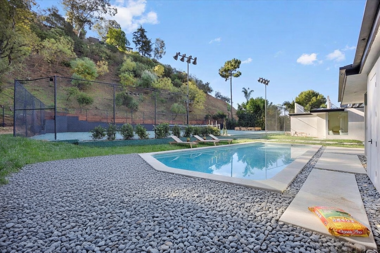 This outdoor space features a rectangular swimming pool set against a backdrop of a hillside and a private tennis court. The area includes a paved walkway, a grassy lawn, and several lounge chairs positioned near the water. The scene captures a serene, resort-like atmosphere perfect for recreation and relaxation.
