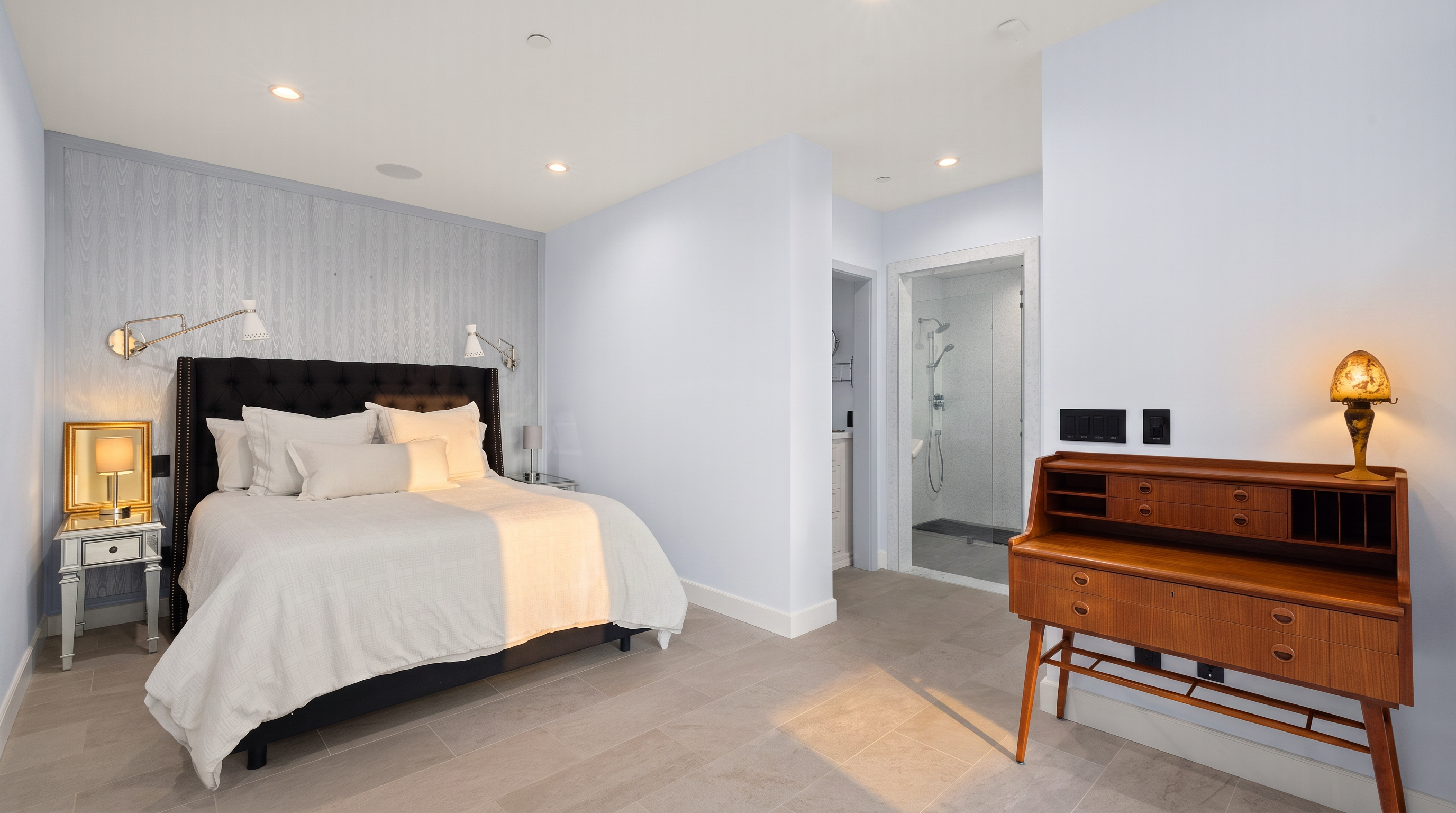 This primary bedroom features a modern aesthetic with light blue walls and a textured accent wall behind a black tufted headboard. The room is furnished with a white-covered bed, mirrored nightstands, and a vintage-style wooden desk in the foreground. A doorway leads to an en-suite bathroom with a glass-enclosed shower, all set against neutral tile flooring.