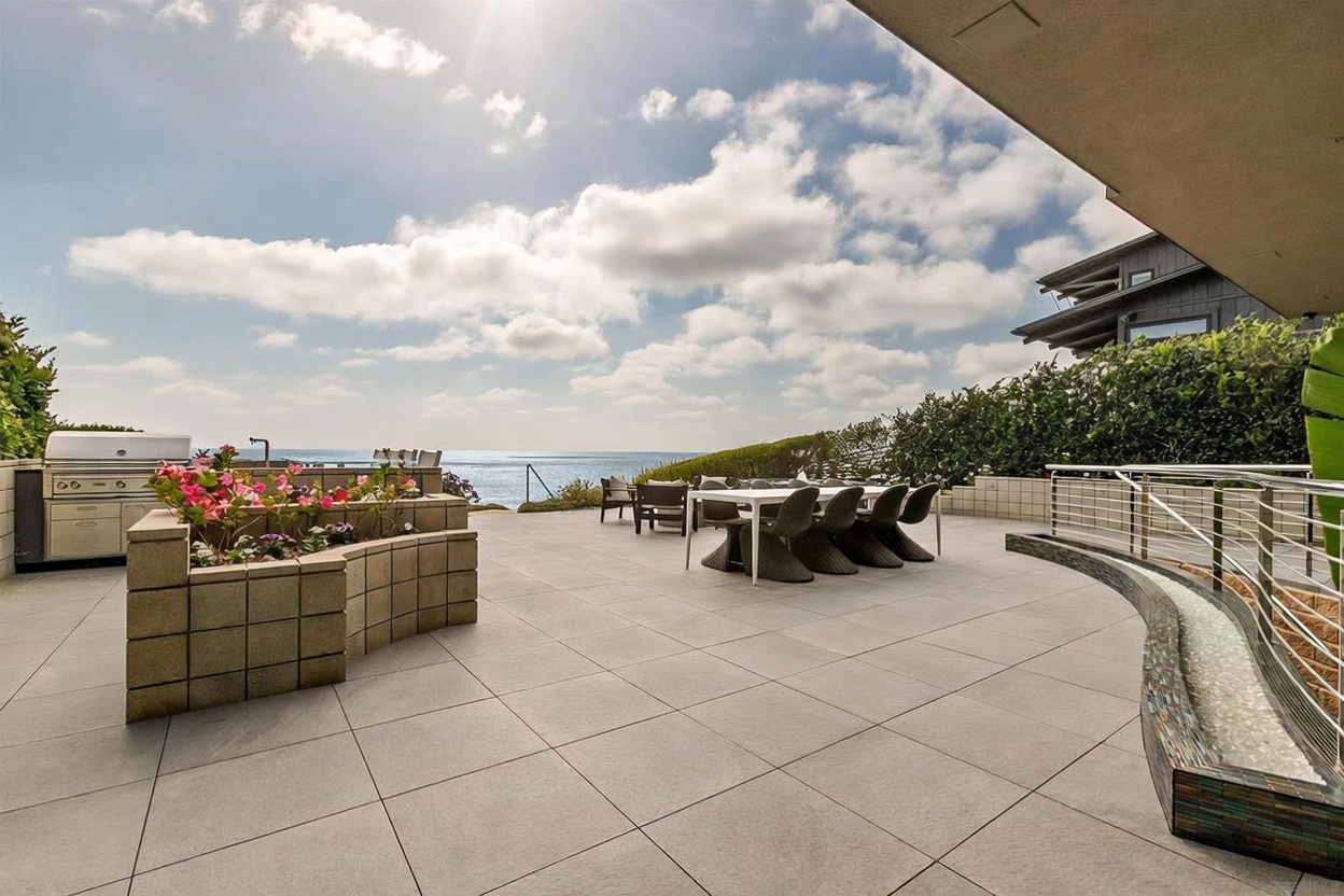 This expansive outdoor patio offers a stunning, elevated view of the ocean under a bright, cloudy sky. The space is well-appointed with a modern dining set, a built-in outdoor kitchen area with a grill, and a curved decorative water feature along the railing. The clean, paved surface and open layout create an ideal setting for coastal entertaining and relaxation.