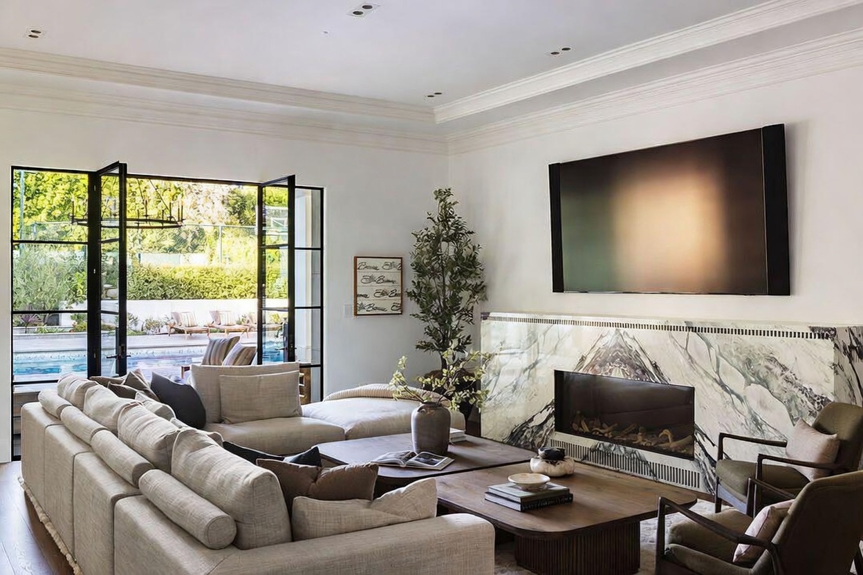 This sophisticated living room features a large, light-toned sectional sofa arranged around two low-profile wooden coffee tables. A striking stone-clad chimney breast serves as the focal point, housing a modern linear fireplace beneath a large wall-mounted television. Large, black-framed glass doors open to reveal a bright outdoor pool area, creating a seamless transition between indoor and outdoor living spaces.