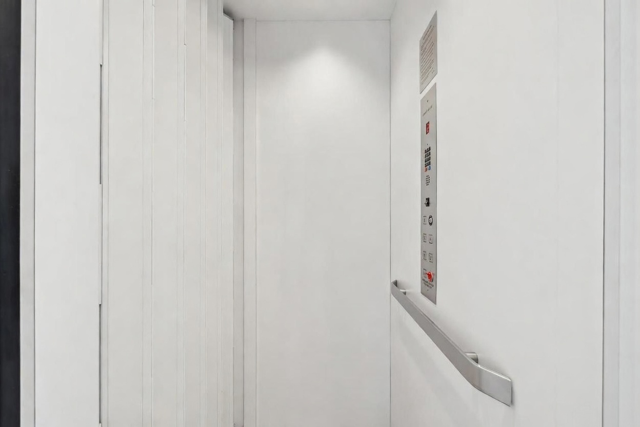 This image shows the interior of a minimalist, modern elevator cab finished in a clean, bright white. A vertical control panel with various floor buttons is mounted on the right wall, accompanied by a sleek, horizontal metal handrail. The perspective is centered, looking directly into the small, enclosed space, emphasizing its simple and functional design.