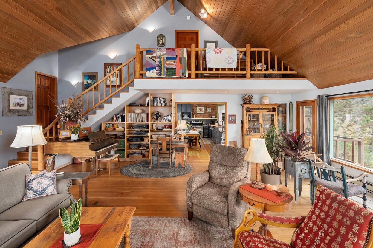 This spacious, open-concept living area features a vaulted wood-paneled ceiling and a cozy, rustic aesthetic. The room includes a grand piano, a comfortable seating area with patterned armchairs, and a built-in shelving unit that leads to a staircase and a lofted landing above. Large windows provide natural light and views of the outdoors, while the warm wood flooring and eclectic decor create a welcoming, homey atmosphere.