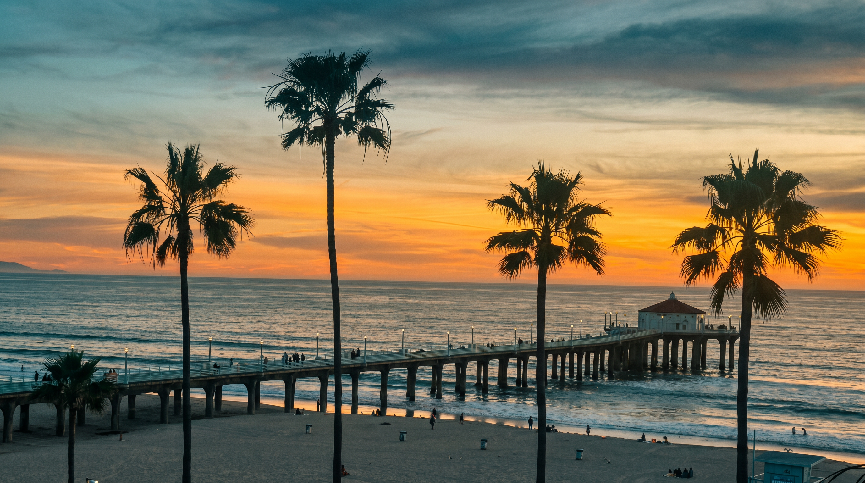 This scenic view captures a sunset over the ocean, featuring a long pier extending into the water with a building at its end. In the foreground, several tall palm trees frame the sandy beach, which is dotted with small figures of people enjoying the evening. The composition offers a cinematic perspective from an elevated vantage point, highlighting the tranquil coastal atmosphere.
