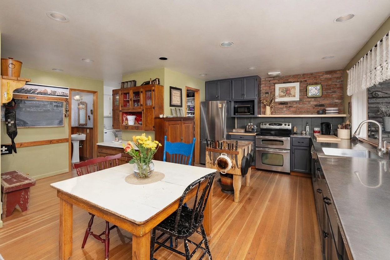This rustic-inspired kitchen features a central dining table with mismatched chairs, a butcher block island, and a distinctive exposed brick wall behind the stainless steel range. The space is accented by dark grey cabinetry, light wood flooring, and a vintage-style wooden hutch, creating a warm and eclectic atmosphere. The perspective is a wide-angle shot from the dining area looking toward the cooking zone, capturing the functional layout and cozy character of the home.