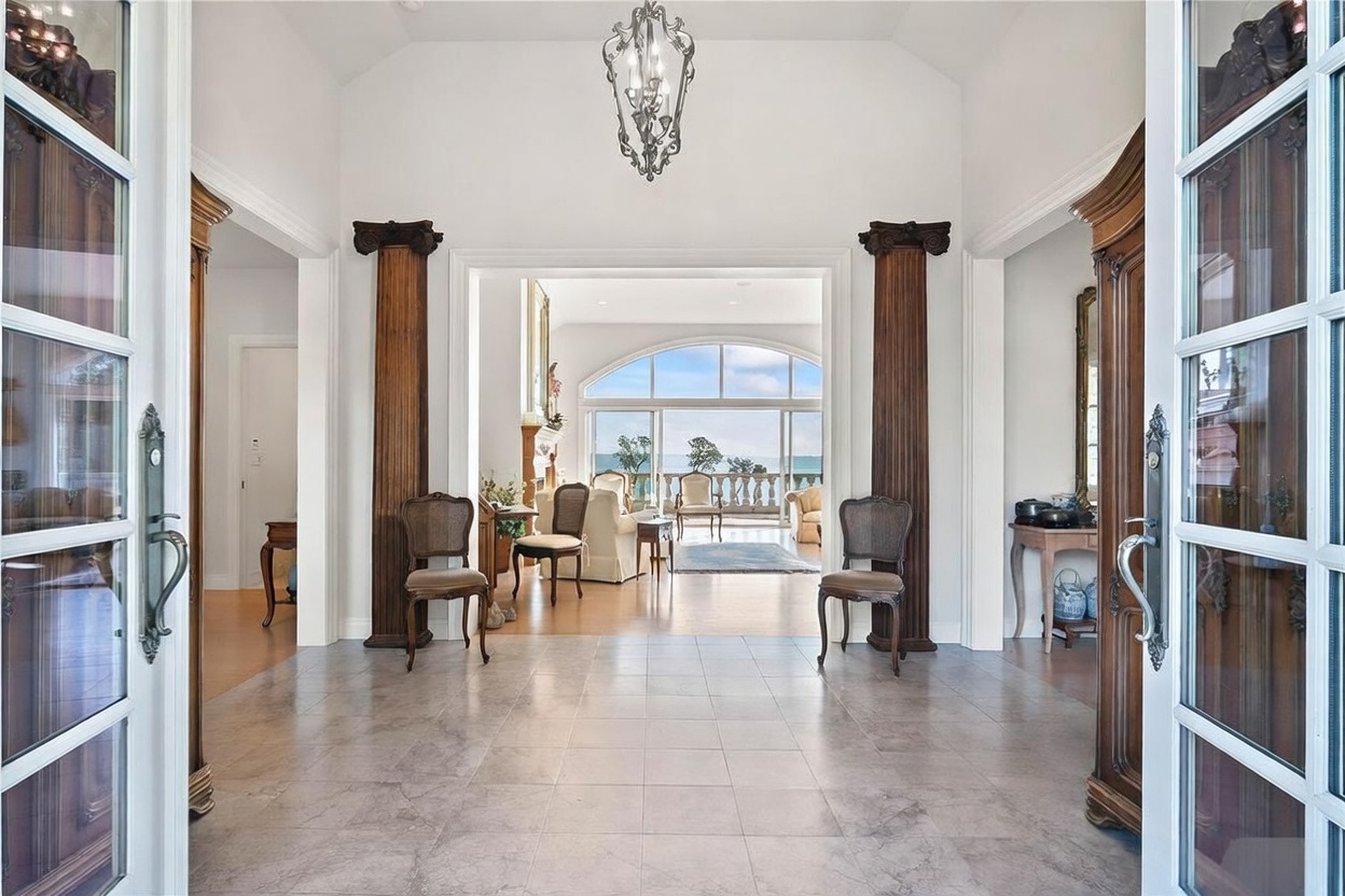 This grand entryway features a tiled floor and a high ceiling accented by an ornate chandelier, creating an elegant and welcoming atmosphere. Two dark wood columns frame the transition into the living area, which showcases large windows offering a scenic view of the outdoors. The perspective is from the front doorway, looking through the foyer into the bright, open living space beyond.