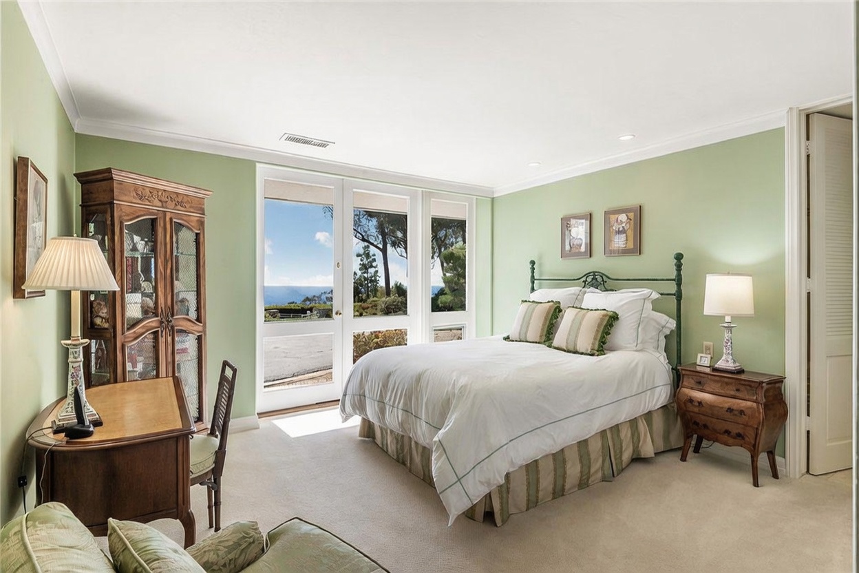 This bright bedroom features light green walls, neutral carpeting, and a queen-sized bed with a dark metal frame. A large wooden armoire and a matching desk provide ample storage and workspace, while tall windows offer a view of the outdoors. The room is accented by two framed pictures above the bed and a traditional bedside table with a lamp, creating a cozy and inviting atmosphere.