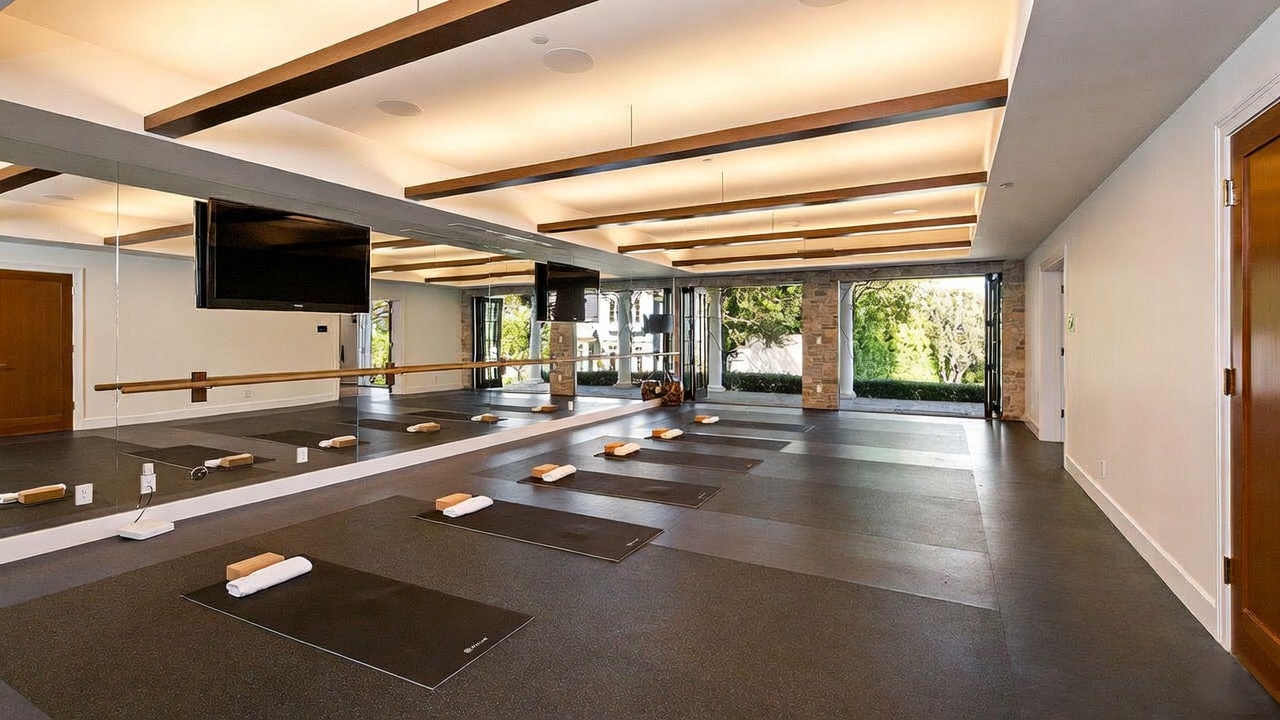 This spacious home gym features a dedicated yoga or workout area with multiple mats laid out on a dark, resilient floor. A long ballet barre is mounted against a wall of mirrors, which reflects the room and the open, folding glass doors that lead to an outdoor patio. The ceiling is accented with decorative wooden beams and recessed lighting, creating a bright and professional atmosphere for fitness activities.