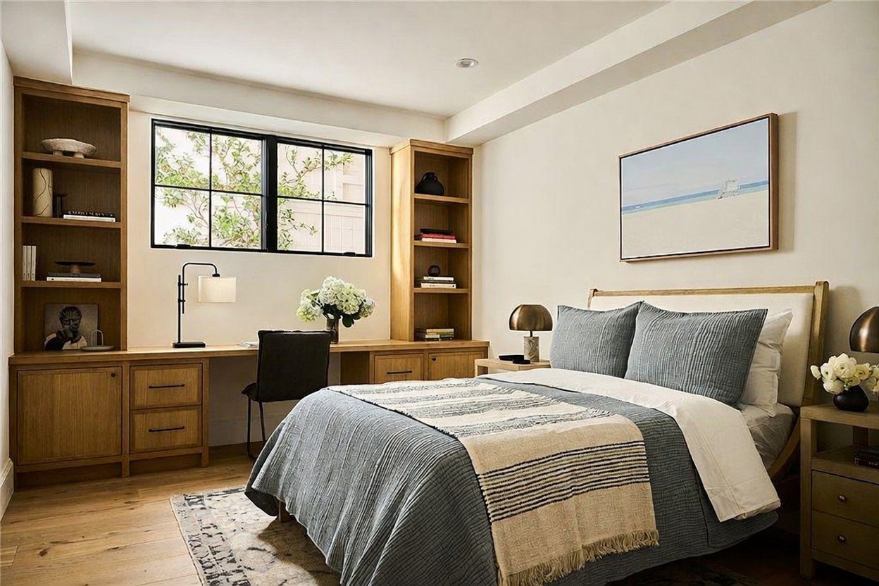 This well-appointed bedroom features a neutral color palette and a built-in desk area flanked by custom light wood shelving. A queen-sized bed with layered blue and white bedding serves as the focal point, complemented by a piece of coastal-themed artwork above the headboard. The room is bathed in natural light from a large window, creating a serene and functional living space.