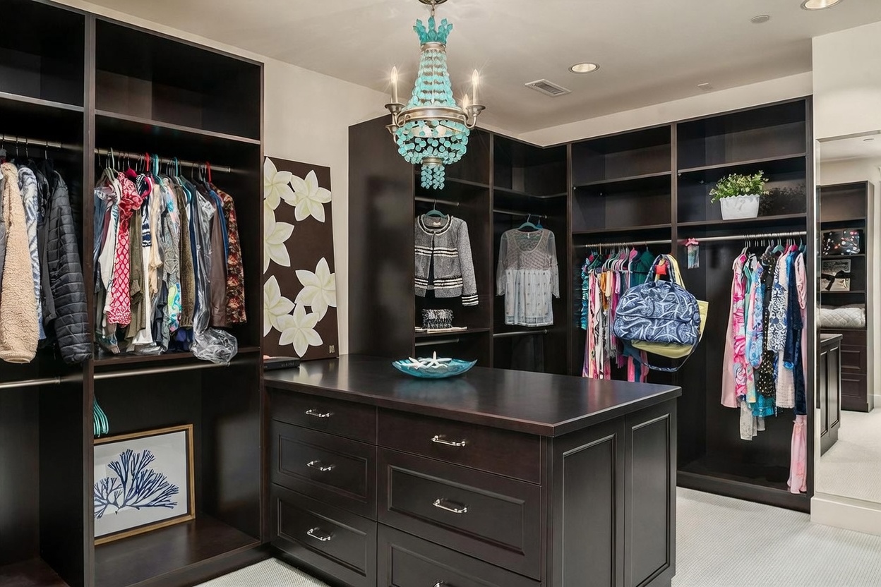 This spacious walk-in closet features dark wood cabinetry with extensive hanging space and shelving, centered around a large island with built-in drawers. A decorative turquoise chandelier serves as a focal point, while the room is accented with coastal-themed artwork and decor. The neutral carpeting and organized layout create a sophisticated and functional dressing area.