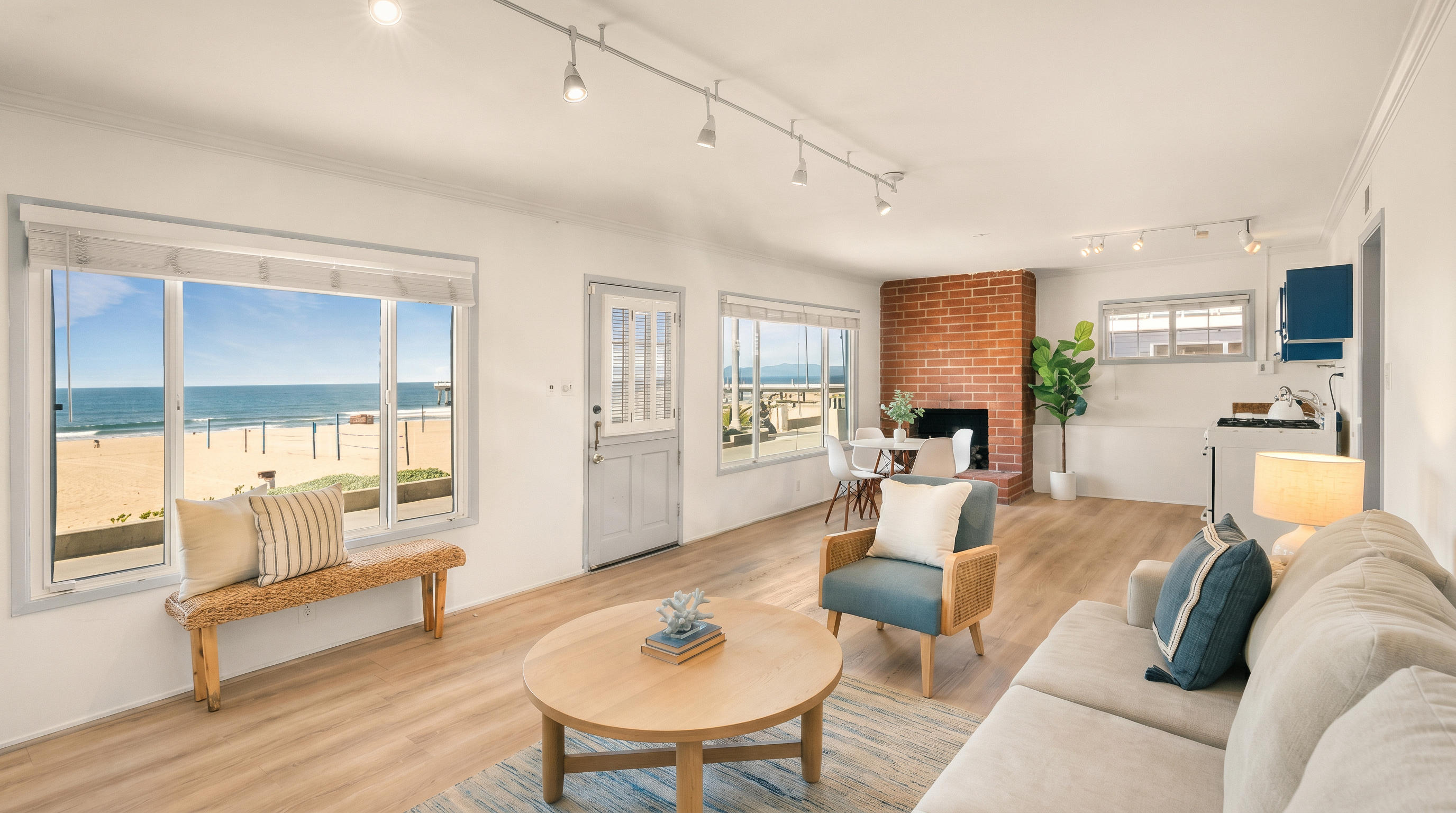This bright and airy living room features light wood-toned flooring and a neutral color palette, emphasizing a relaxed, coastal aesthetic. A prominent brick chimney breast serves as a focal point, while large windows provide expansive, unobstructed views of the beach and ocean. The space is furnished with a comfortable sofa, a round coffee table, and an accent chair, creating an inviting atmosphere that seamlessly blends indoor living with the outdoor scenery.