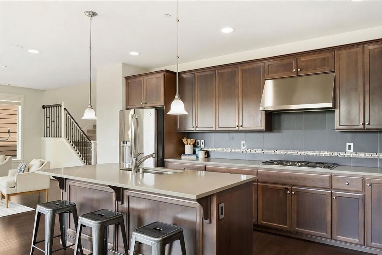 This kitchen features dark wood cabinetry, a large central island with a stone countertop, and stainless steel appliances, including a range hood and refrigerator. The space is illuminated by two pendant lights hanging over the island and recessed ceiling lighting, creating a warm and functional atmosphere. In the background, a staircase with a black metal railing leads to an upper level, while a glimpse of a living area with a light-colored sofa is visible to the left.