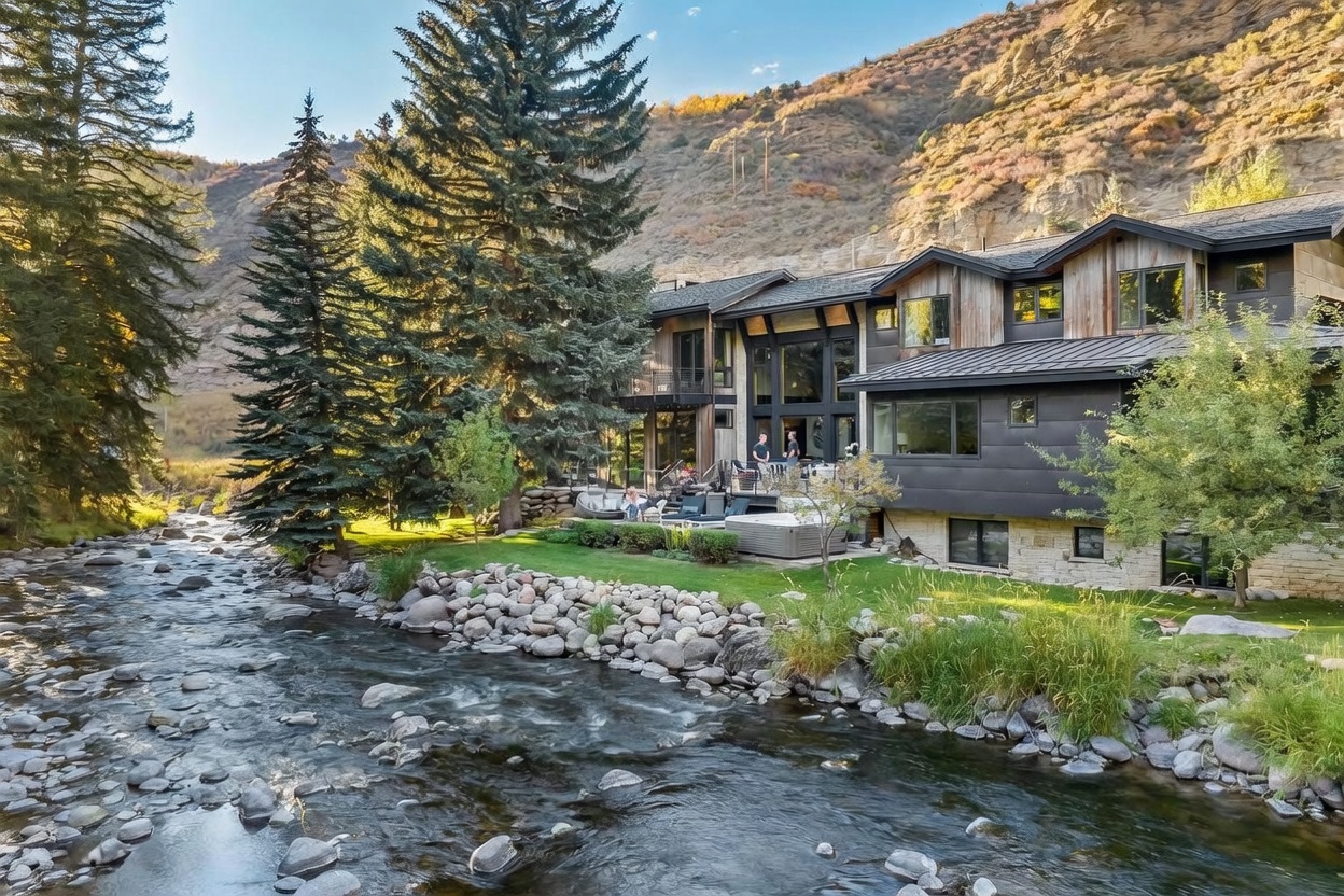 This stunning mountain residence features a modern architectural design with a mix of dark metal siding and natural wood accents, situated directly along a rushing river. The rear exterior includes a spacious multi-level deck with outdoor seating and a hot tub, seamlessly blending the home with the surrounding forest and rocky landscape. The perspective captures the home's integration into the rugged terrain, emphasizing the tranquil waterfront setting and expansive glass windows that connect the interior to the natural environment.