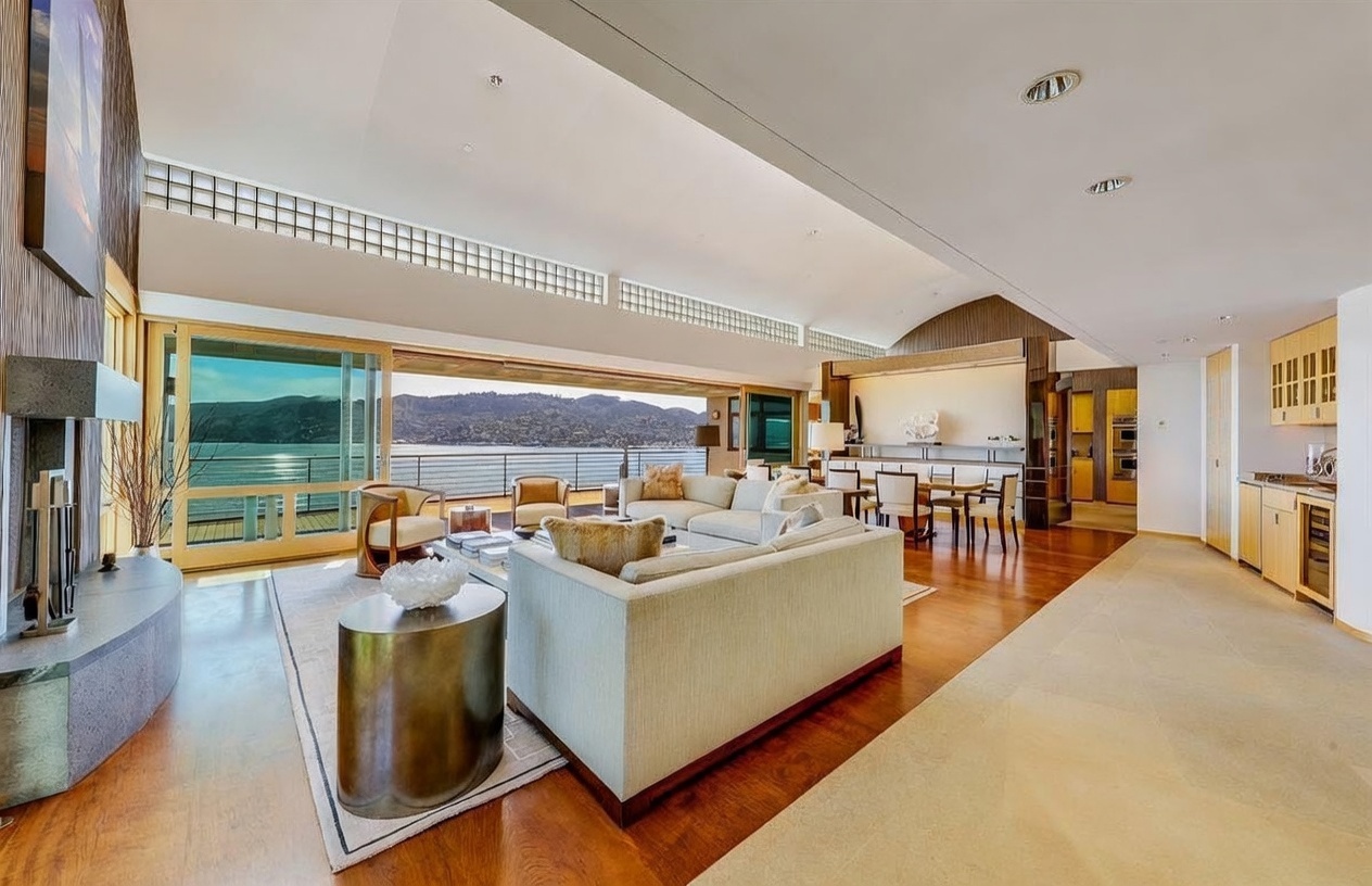 This expansive, open-concept living area features a sophisticated contemporary design with warm wood flooring and a neutral color palette. A large, light-colored sectional sofa anchors the space, which flows seamlessly into a dining area and a kitchen with light wood cabinetry. The room is defined by a massive wall of glass that opens to a deck, providing panoramic views of the water and distant hills, while a tall, stone-clad chimney breast adds a textured focal point to the left.