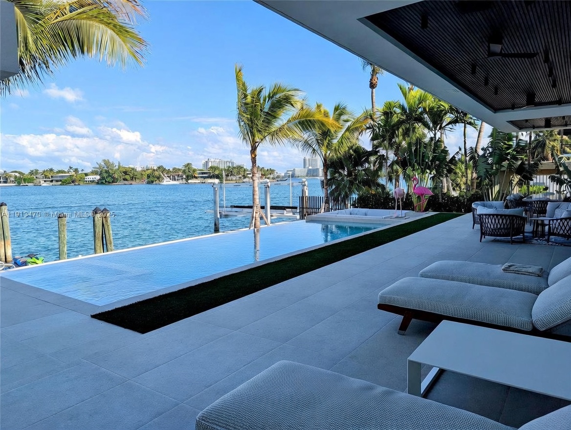 This luxurious waterfront patio features a sleek infinity-edge pool that seamlessly blends with the bay beyond. The expansive deck is furnished with comfortable lounge chairs, creating an ideal space for relaxation while overlooking the calm water and distant city skyline. Lush palm trees and tropical landscaping frame the view, enhancing the serene and high-end atmosphere of this outdoor living area.