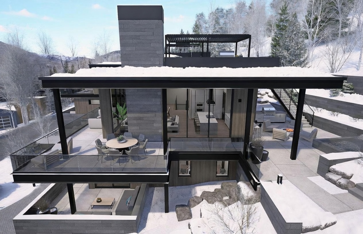 This aerial perspective captures a modern, multi-level mountain residence set against a snowy, forested landscape. The home features a prominent stone-clad chimney breast, expansive glass walls that open to a spacious deck with outdoor dining, and a lower-level patio area. The architectural design emphasizes clean lines, dark structural elements, and seamless indoor-outdoor living, perfectly suited for a luxury alpine retreat.