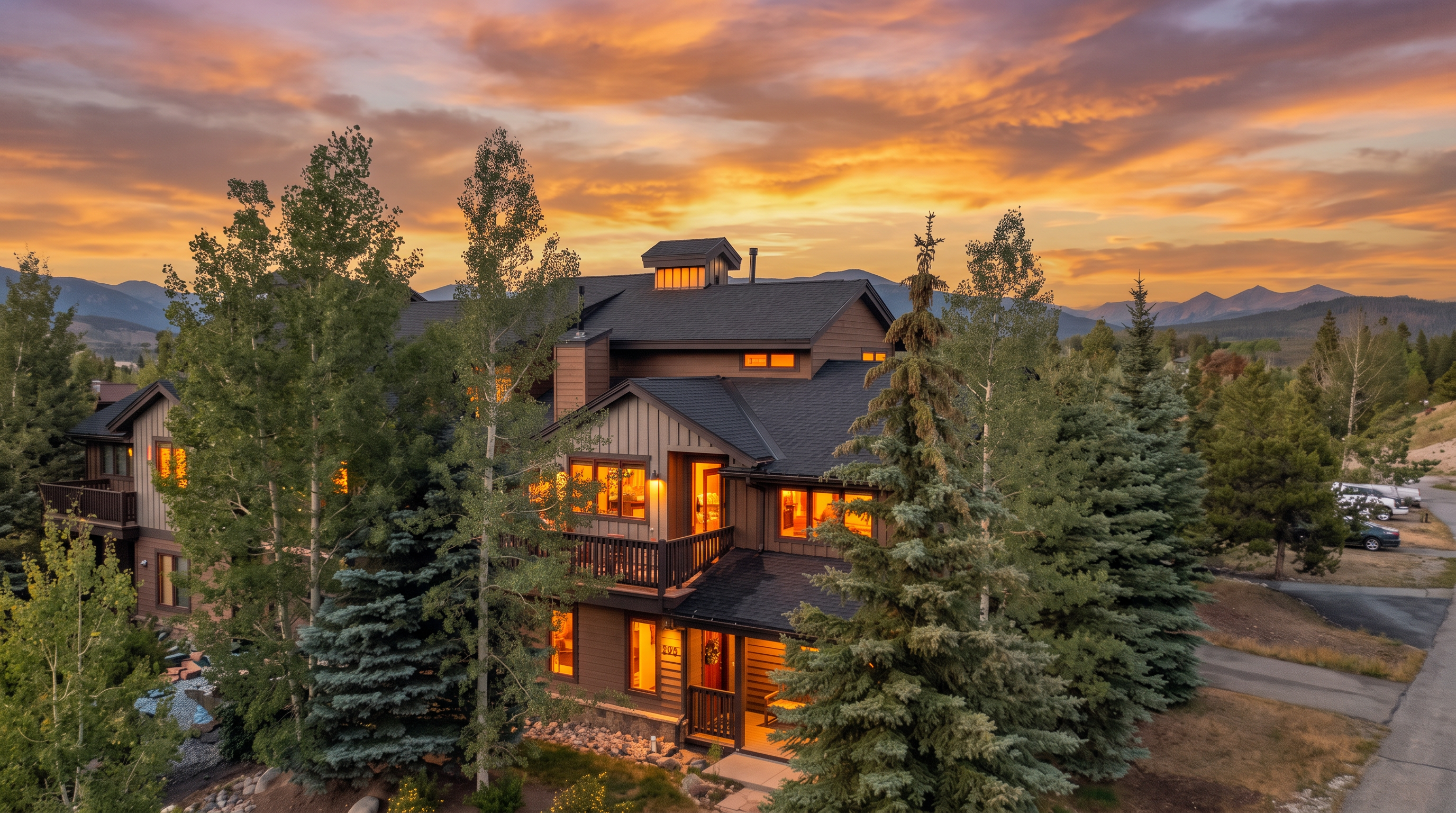 This elevated aerial perspective captures a multi-story mountain home nestled among mature pine and aspen trees. The structure features a dark shingled roof, brown vertical siding, and a prominent balcony overlooking the entrance, with a stone-accented foundation. The warm glow from the interior windows contrasts beautifully with the soft, golden light of the sunset sky in the background.