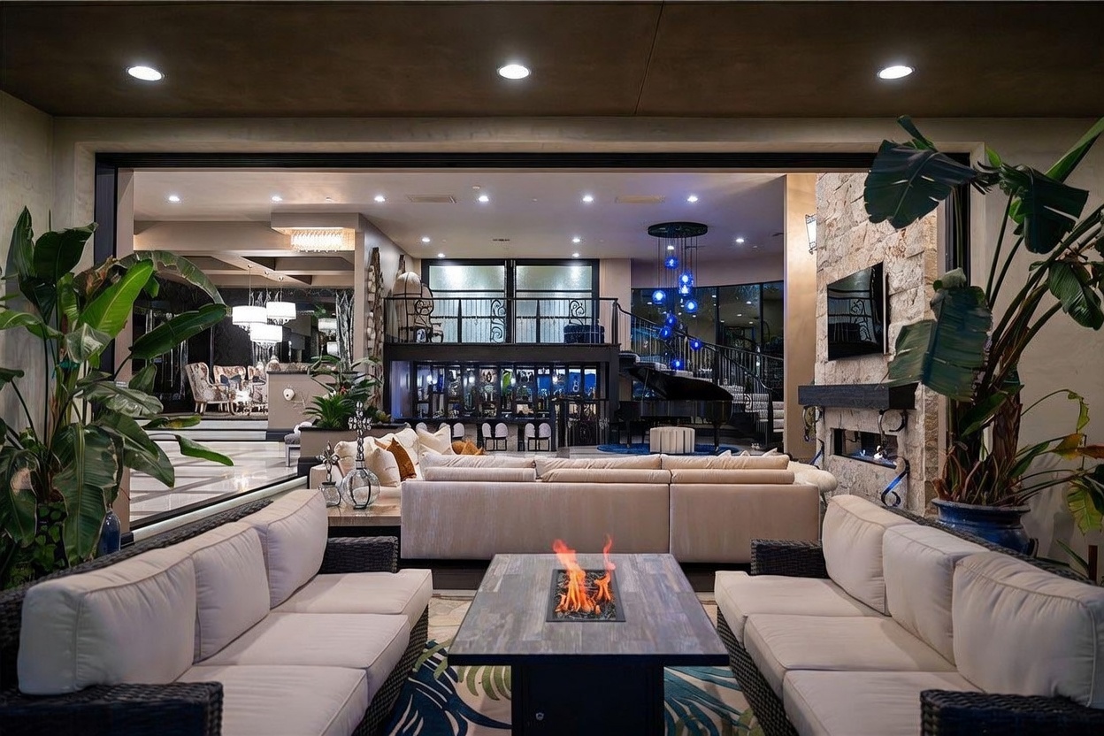 This luxurious outdoor living space features a large, covered patio area seamlessly connected to an expansive indoor living room through a wide, open-concept transition. The patio is furnished with comfortable, light-toned lounge seating arranged around a central fire table, while the interior showcases a sophisticated bar area, a grand piano, and elegant blue pendant lighting. The overall aesthetic is one of high-end, resort-style living, blending indoor comfort with an open-air atmosphere.
