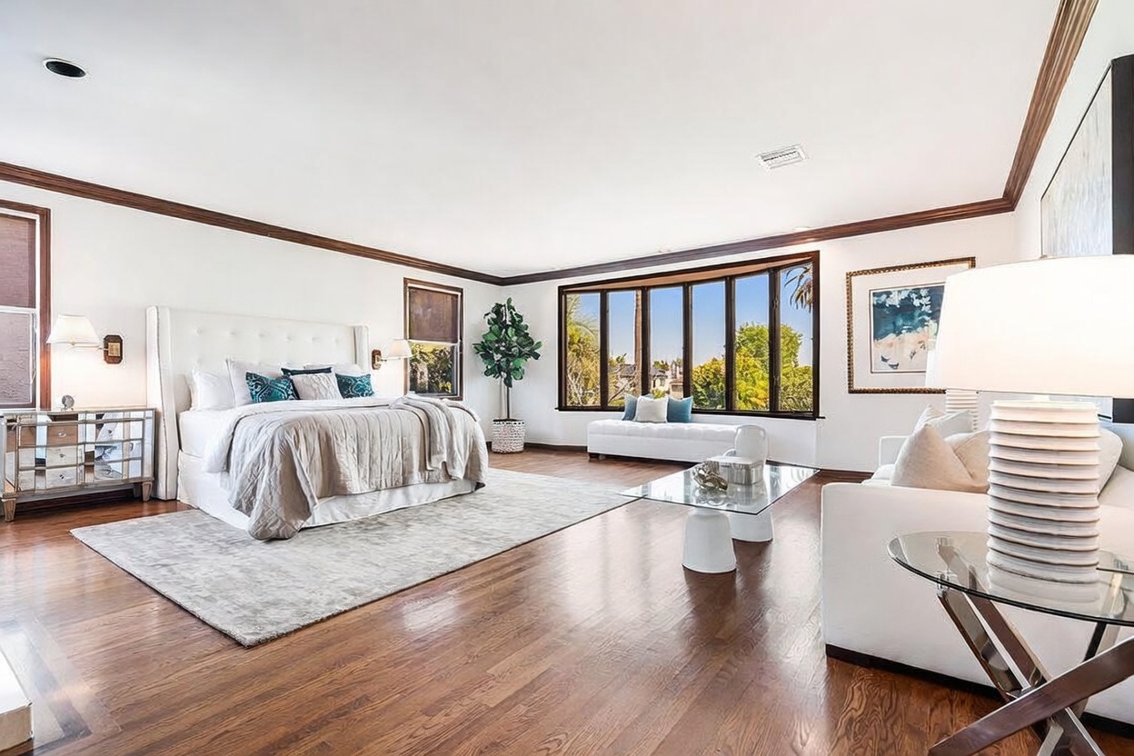 This spacious primary bedroom features rich hardwood flooring and a neutral color palette, centered around a large tufted headboard. A seating area with a white sofa and a glass-topped coffee table sits to the right, while a wall of large windows provides ample natural light and views of the outdoors. The room is accented by a potted plant, a mirrored nightstand, and a framed piece of art, creating a bright and airy atmosphere.