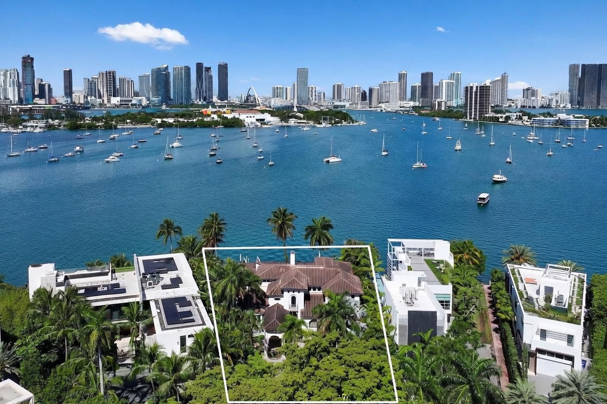 This high-angle aerial view captures a waterfront residential property highlighted by a white outline, nestled among lush tropical landscaping. The home features a traditional tiled roof and is flanked by modern, flat-roofed residences, all situated along a vibrant blue bay filled with numerous sailboats. In the background, a sprawling city skyline rises against a clear blue sky, providing a dramatic urban backdrop to this serene coastal setting.