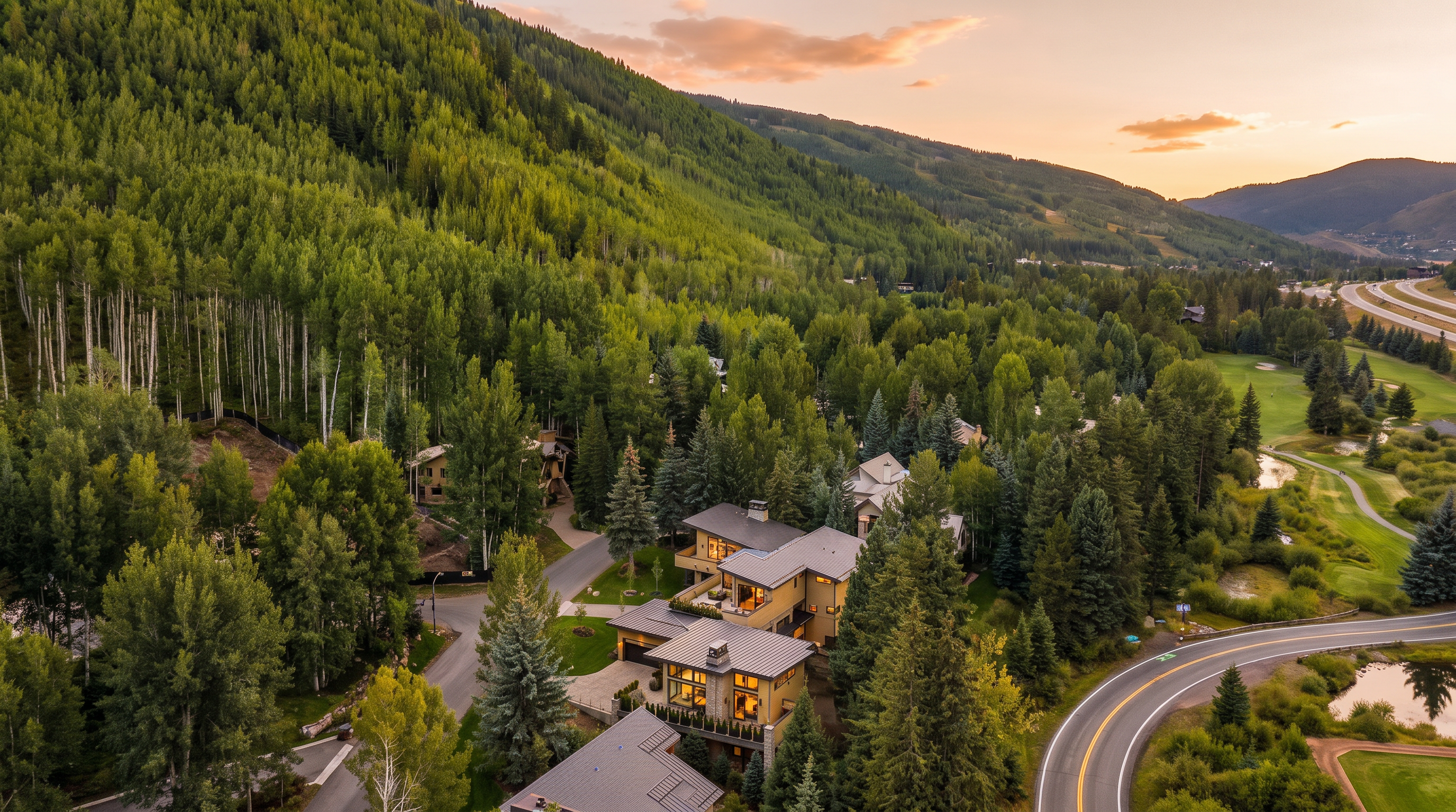 This high-angle aerial view captures a contemporary mountain residence nestled within a dense forest of aspen and pine trees. The multi-level home features a modern architectural design with dark metal roofing, stone accents, and expansive decks that overlook the surrounding landscape and a nearby golf course. A winding road curves past the property, emphasizing its secluded yet accessible location in a scenic, mountainous environment.
