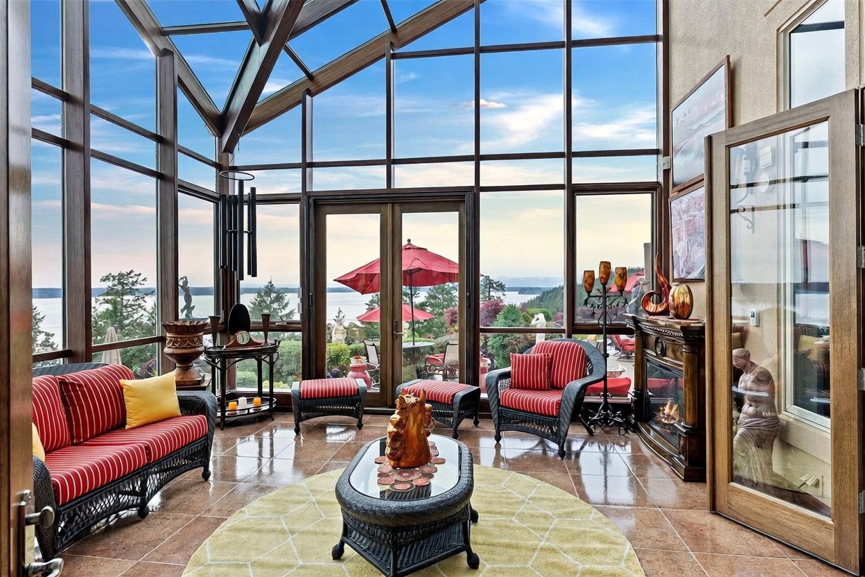 This elegant sunroom features expansive floor-to-ceiling glass walls and a vaulted ceiling, offering panoramic views of the water and surrounding landscape. The space is furnished with a matching set of dark wicker seating with red-and-white striped cushions, centered around a glass-topped coffee table on a circular yellow rug. A traditional fireplace with a wooden mantel adds warmth to the room, which is finished with polished tile flooring and decorative accents.