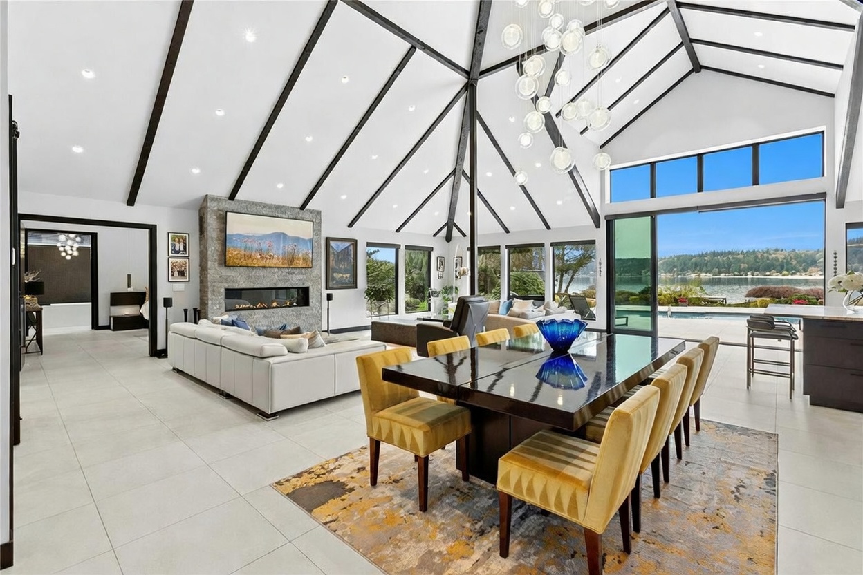 This expansive, open-concept living and dining area features a dramatic vaulted ceiling with exposed dark beams and a striking modern chandelier. A floor-to-ceiling stone fireplace serves as the focal point, while large glass sliding doors provide a seamless transition to a scenic waterfront patio. The space is elegantly furnished with a contemporary white sectional and a sophisticated dining set, creating a bright, airy, and luxurious atmosphere.