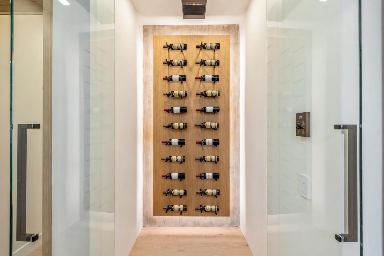 This sophisticated wine cellar features a central light wood display panel mounted on a white wall, showcasing two vertical columns of wine bottles stored horizontally. The space is framed by glass doors on either side, creating a modern and transparent aesthetic. A digital climate control panel is mounted on the right wall, emphasizing the functional, high-end nature of this storage area.