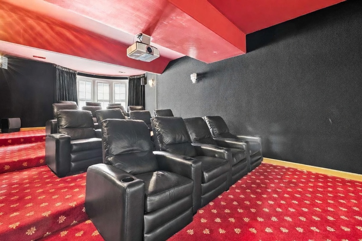 This dedicated home theater room features tiered seating with multiple rows of black leather recliners facing a projection screen area. The space is designed with a bold aesthetic, utilizing a vibrant red carpet with a repeating pattern and a matching red ceiling, contrasted by dark, textured walls. A ceiling-mounted projector is positioned above the seating area, and a bay window with blinds is visible at the back of the room.