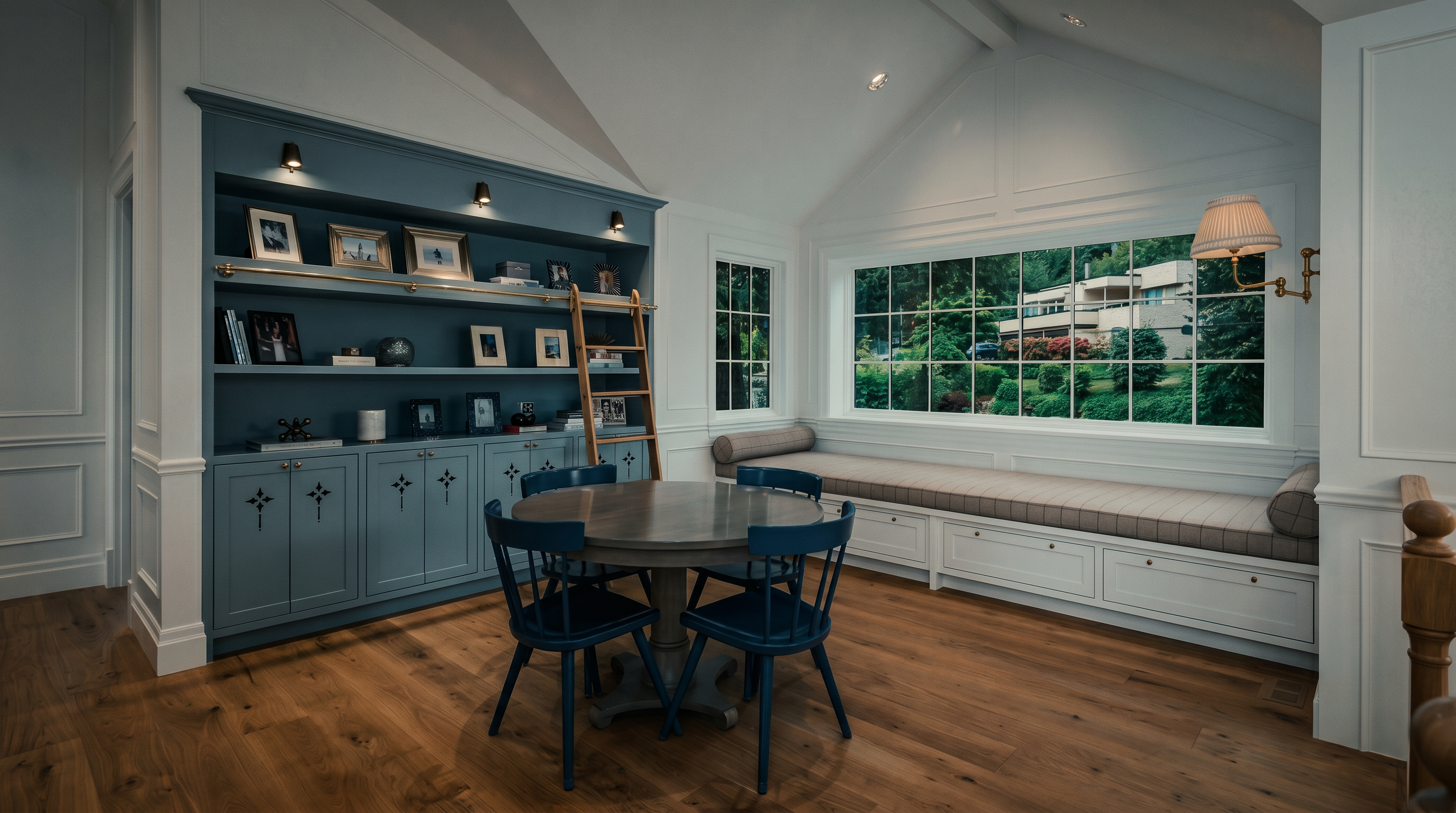 This bright and airy study features a vaulted ceiling and light wood flooring, creating a welcoming atmosphere. A large, built-in blue shelving unit with an integrated rolling ladder serves as a focal point, complemented by a central round table and blue chairs. A long window seat built into a wide window frame offers a comfortable nook with views of the lush greenery outside.