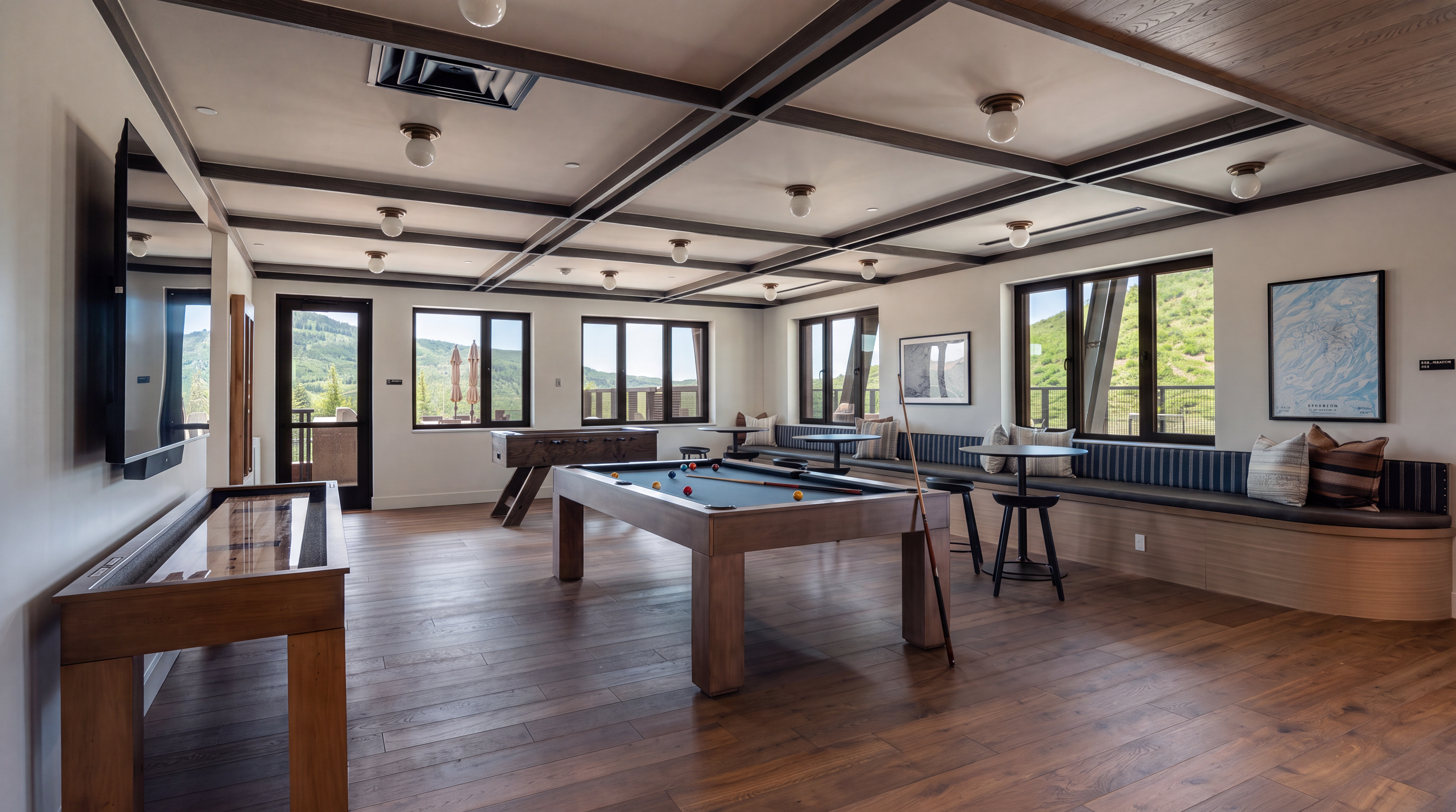 This spacious game room features a pool table as the central focal point, complemented by a shuffleboard table and a foosball table. The room is designed with a long, built-in bench seating area along the windows, which offer scenic views of the surrounding landscape. The space is finished with warm wood flooring and a coffered ceiling with recessed lighting, creating an inviting atmosphere for recreation.