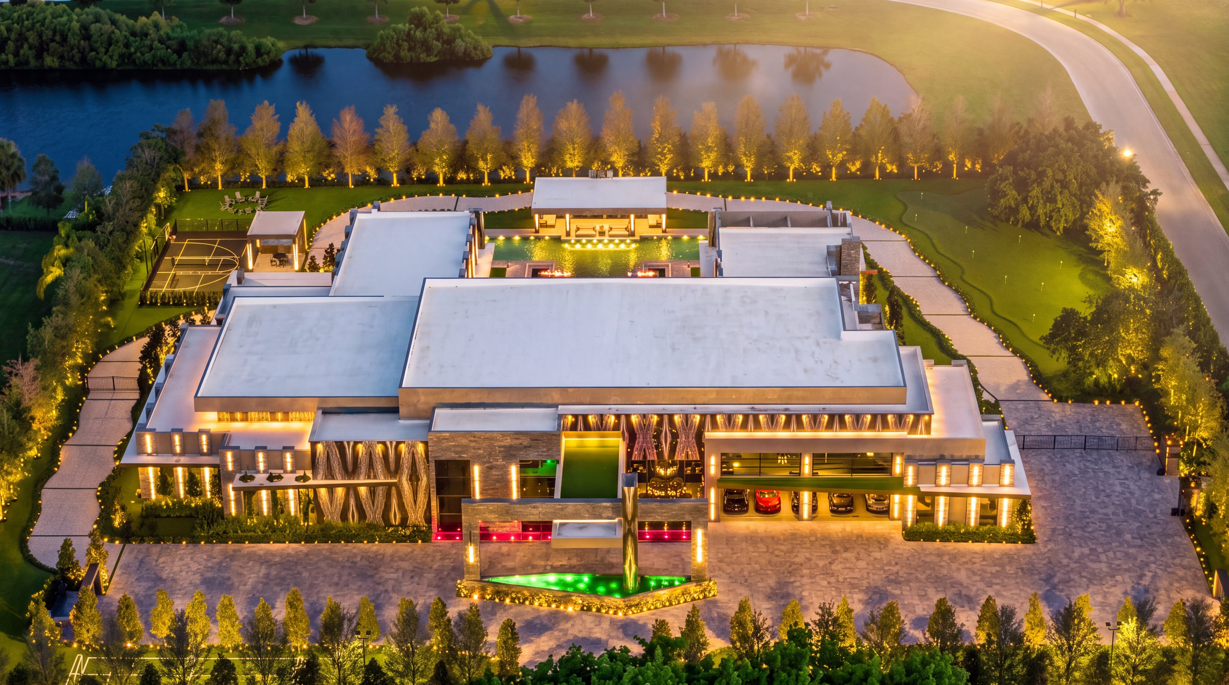 This stunning aerial perspective captures a sprawling, modern luxury estate illuminated at dusk. The property features a massive central structure with a large swimming pool, a private basketball court, and a manicured putting green, all set against a backdrop of a serene lake and lush landscaping. The architectural design emphasizes symmetry and grand scale, with integrated lighting highlighting the driveway, water features, and the expansive garage area.