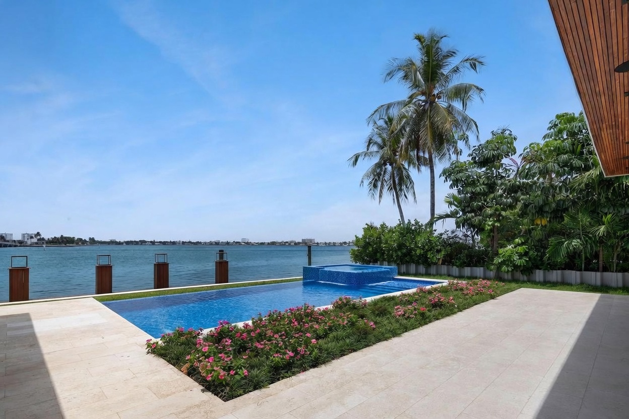 This luxurious waterfront property features a sleek, rectangular infinity-edge pool with an integrated raised spa. The pool deck is paved with light-toned stone, bordered by a vibrant garden bed filled with pink flowering shrubs and lush greenery. Beyond the pool, the expansive view showcases a calm bay with distant buildings, creating a serene and high-end outdoor living space.
