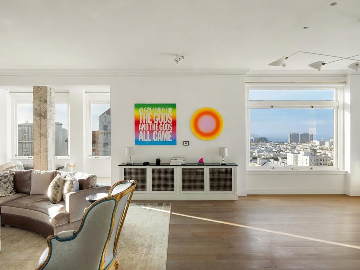 This bright and airy living space features an open-concept layout with light wood flooring and large windows that frame a panoramic city view. The room is anchored by a plush, neutral-toned sectional sofa and a long console table topped with modern art and lamps. The aesthetic is contemporary and sophisticated, highlighted by vibrant wall art and a minimalist dining area to the right.