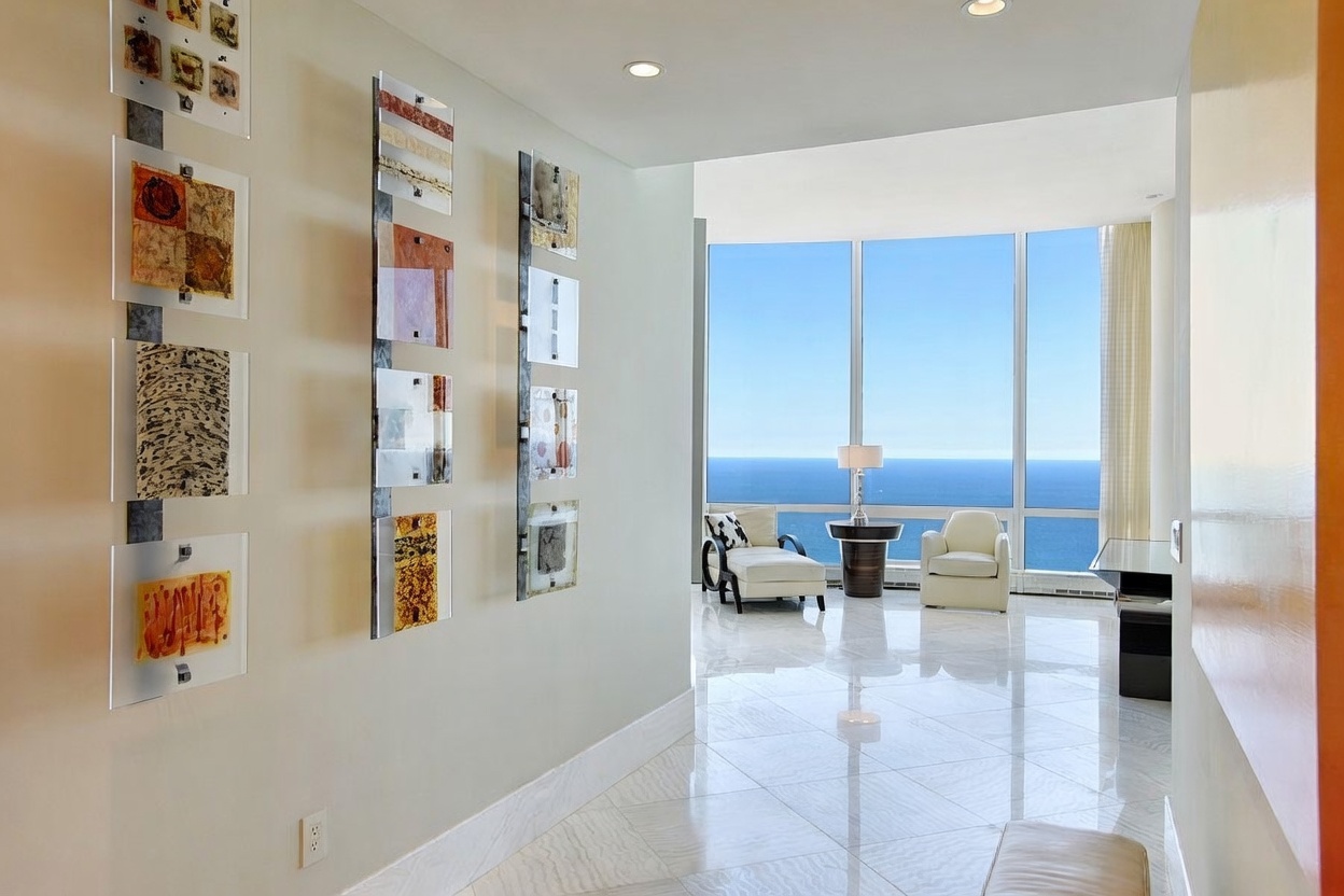 This perspective captures a bright hallway leading into a spacious living area with expansive floor-to-ceiling windows overlooking the ocean. The hallway walls are adorned with a series of decorative glass art panels, while the floor features light-toned, large-format tile. The transition from the gallery-like corridor to the open, sun-drenched living space creates an airy and sophisticated atmosphere.