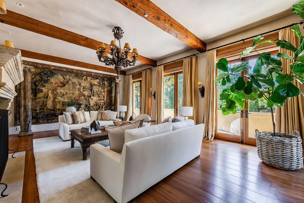 This elegant living room features exposed wooden ceiling beams, a large decorative tapestry mounted on the wall, and a classic chandelier. The space is furnished with two white sofas arranged around a dark wood coffee table, all resting on a light-colored area rug. Large glass doors with tall curtains allow natural light to fill the room, while a potted plant in a woven basket adds a touch of greenery to the corner.