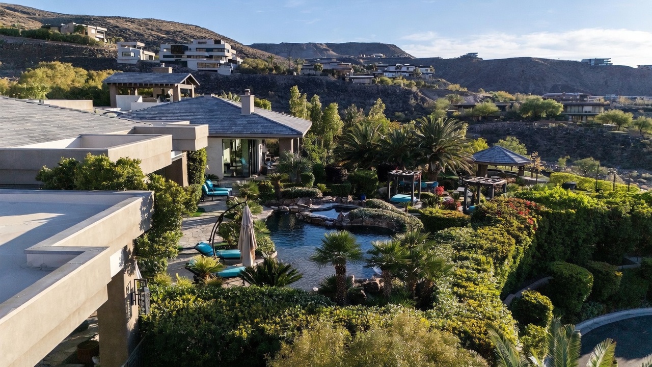 This elevated view captures a lush, landscaped backyard featuring a free-form swimming pool and integrated spa surrounded by mature palm trees and manicured hedges. The property includes a stone-paved patio area with lounge seating and a shaded gazebo, all set against a backdrop of arid hills and neighboring luxury homes. The scene conveys a serene, resort-like atmosphere perfect for outdoor entertaining.