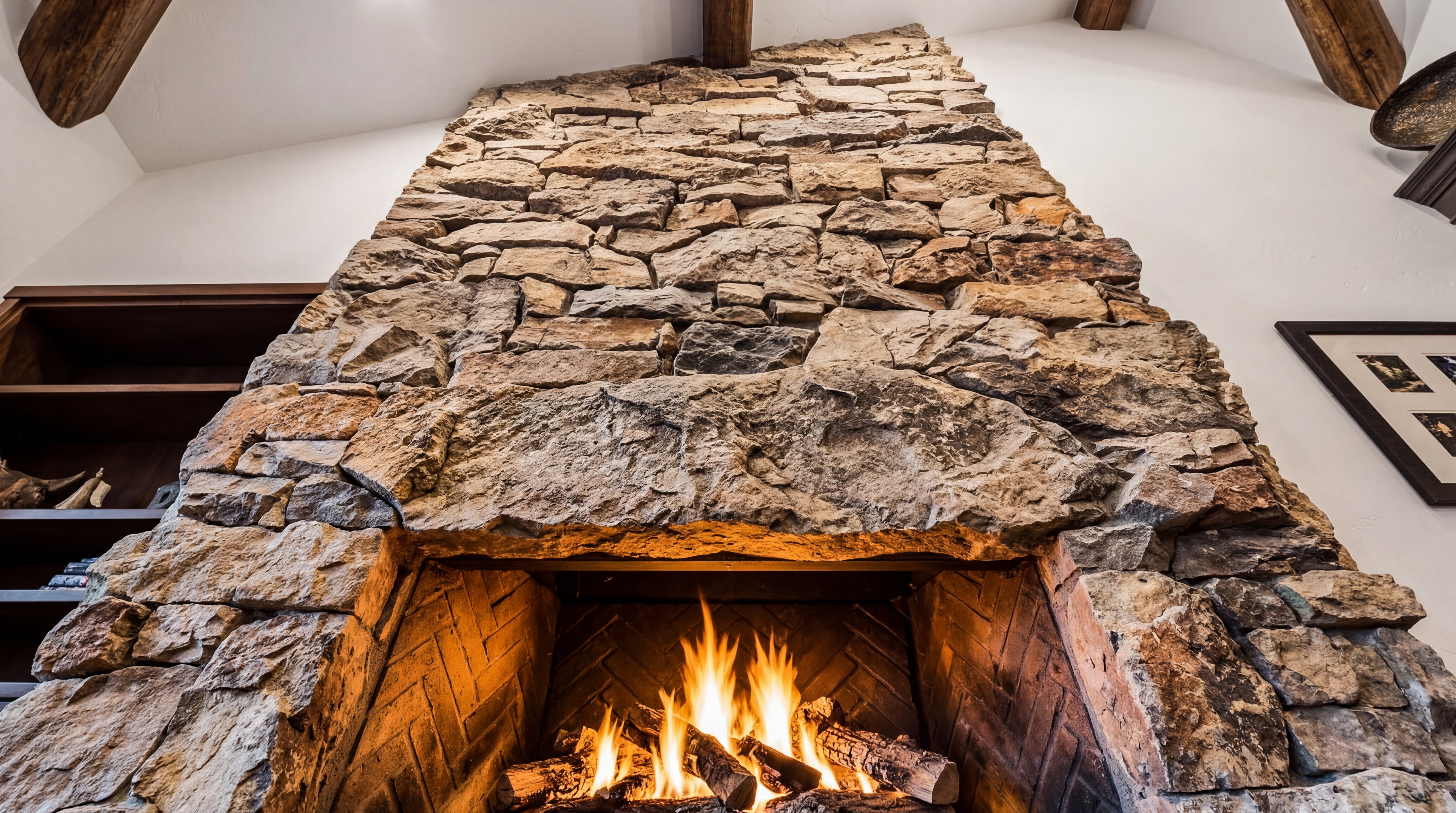 This stunning floor-to-ceiling fireplace serves as a dramatic architectural focal point, crafted from rustic, textured natural stone. The herringbone brick firebox interior complements the rugged exterior stonework, while exposed wooden beams above add warmth and character. The overall composition highlights a high-end, cozy lodge aesthetic.