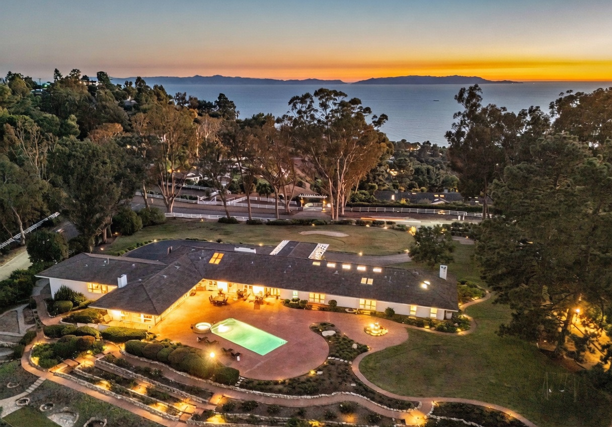 This high-angle aerial view captures a sprawling single-story residence at dusk, set against a dramatic sunset over the ocean. The property features a large, illuminated swimming pool and expansive paved patio area, surrounded by manicured lawns and mature trees. The perspective highlights the home's impressive scale and its prime location overlooking the coastline.