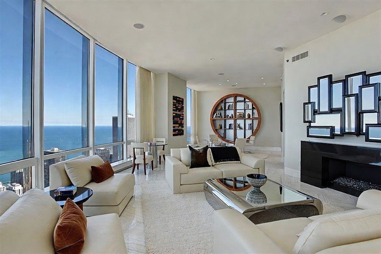 This expansive, high-rise living room features a sophisticated, contemporary design with a neutral color palette and polished stone flooring. The space is defined by its dramatic floor-to-ceiling windows that offer panoramic views of the city skyline and the lake, while a unique circular shelving unit serves as a striking architectural focal point. A sleek, black fireplace surround and a modern mirrored coffee table anchor the seating area, creating an atmosphere of refined luxury and urban elegance.