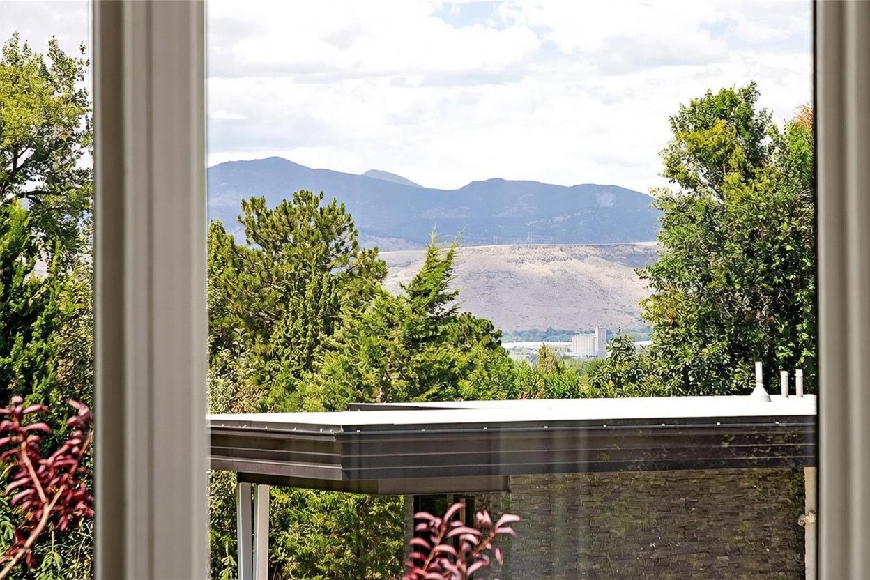 This image is captured from inside looking out through a window, framing a scenic view of distant mountains and a valley. In the foreground, the flat roof of a modern structure with dark horizontal siding is visible, partially obscured by lush green trees. The perspective emphasizes the connection between the interior living space and the expansive natural landscape beyond.