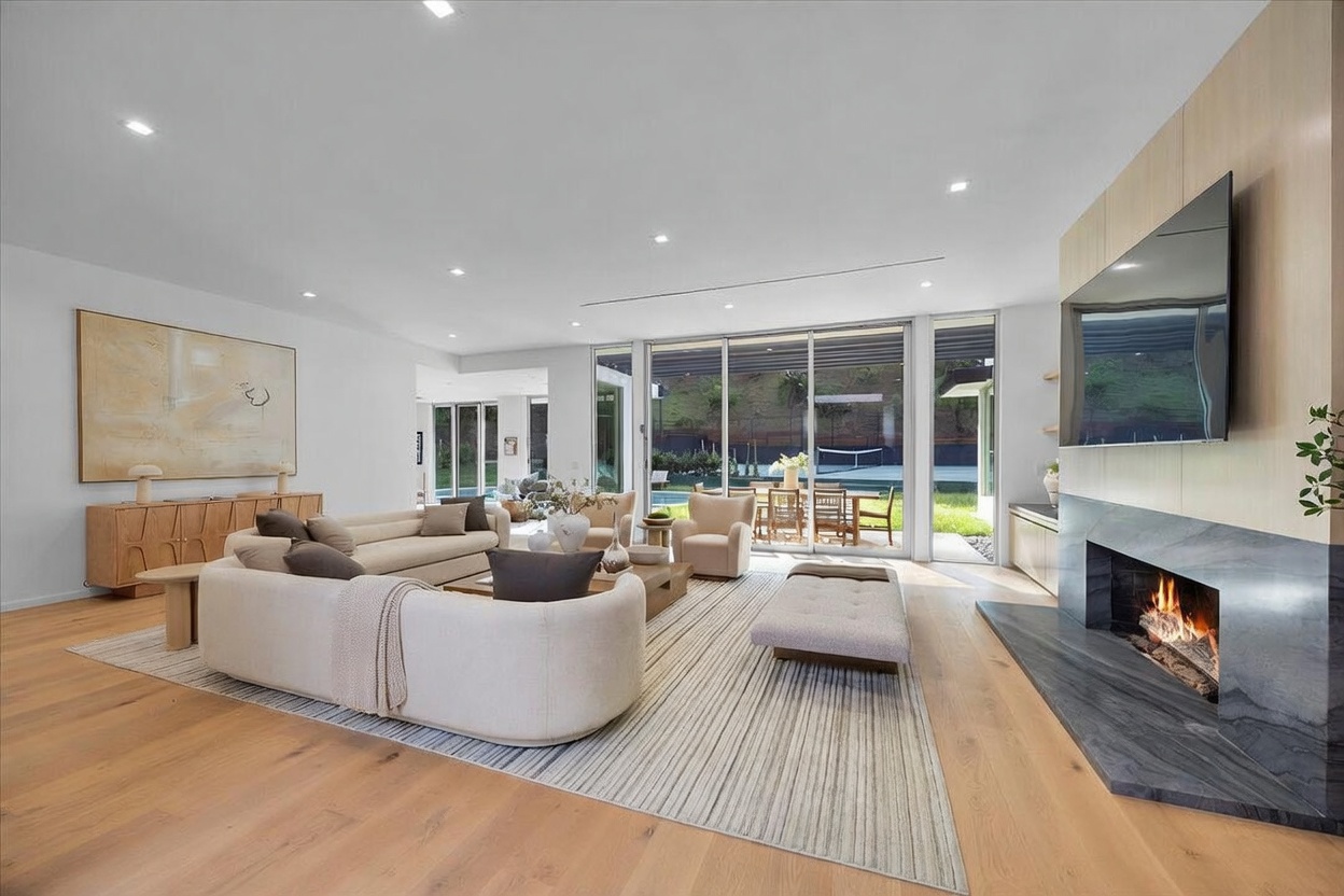 This spacious, open-concept living room features light wood flooring and a neutral color palette, creating an airy and inviting atmosphere. A large, curved sofa and matching armchairs are arranged around a central coffee table, facing a striking fireplace with a dark stone surround and a mounted television. Large glass sliding doors provide a seamless transition to the outdoor patio, allowing for abundant natural light and views of the landscaped yard.
