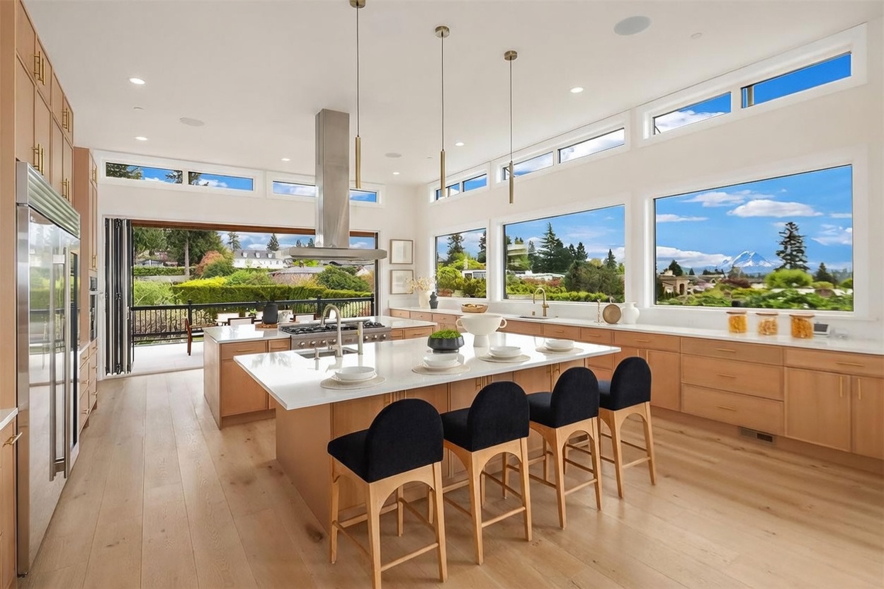 This bright, modern kitchen features light wood cabinetry, a large central island with seating for four, and a professional-grade range. The space is defined by its expansive windows that offer panoramic views of the surrounding landscape and a distant mountain peak. A seamless transition to an outdoor deck is provided by a folding glass door system, creating an airy and open atmosphere.