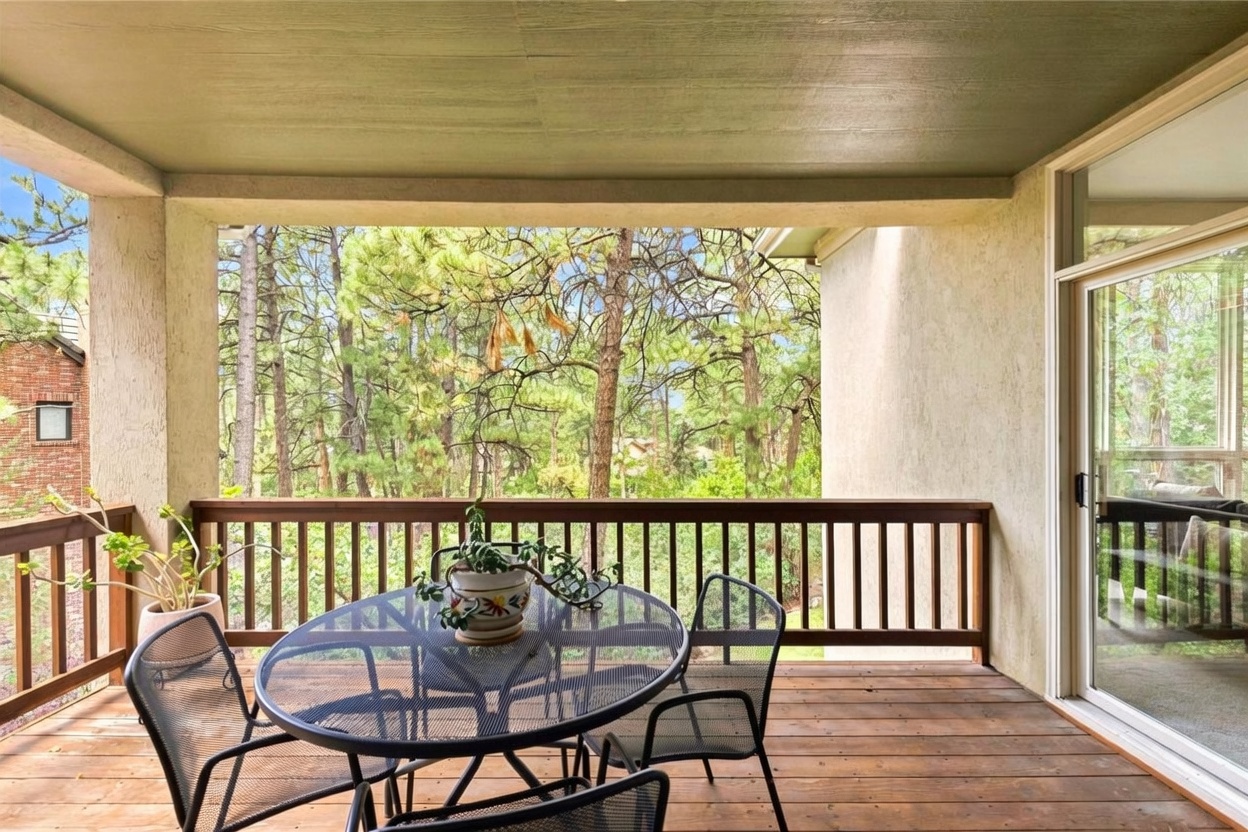 This covered balcony features wooden decking and a simple wooden railing that overlooks a dense, lush forest of pine trees. A metal patio set with a glass-topped table and chairs provides a comfortable outdoor seating area, while a large sliding glass door connects the space to the interior. The setting offers a peaceful, private atmosphere with a scenic, natural backdrop.