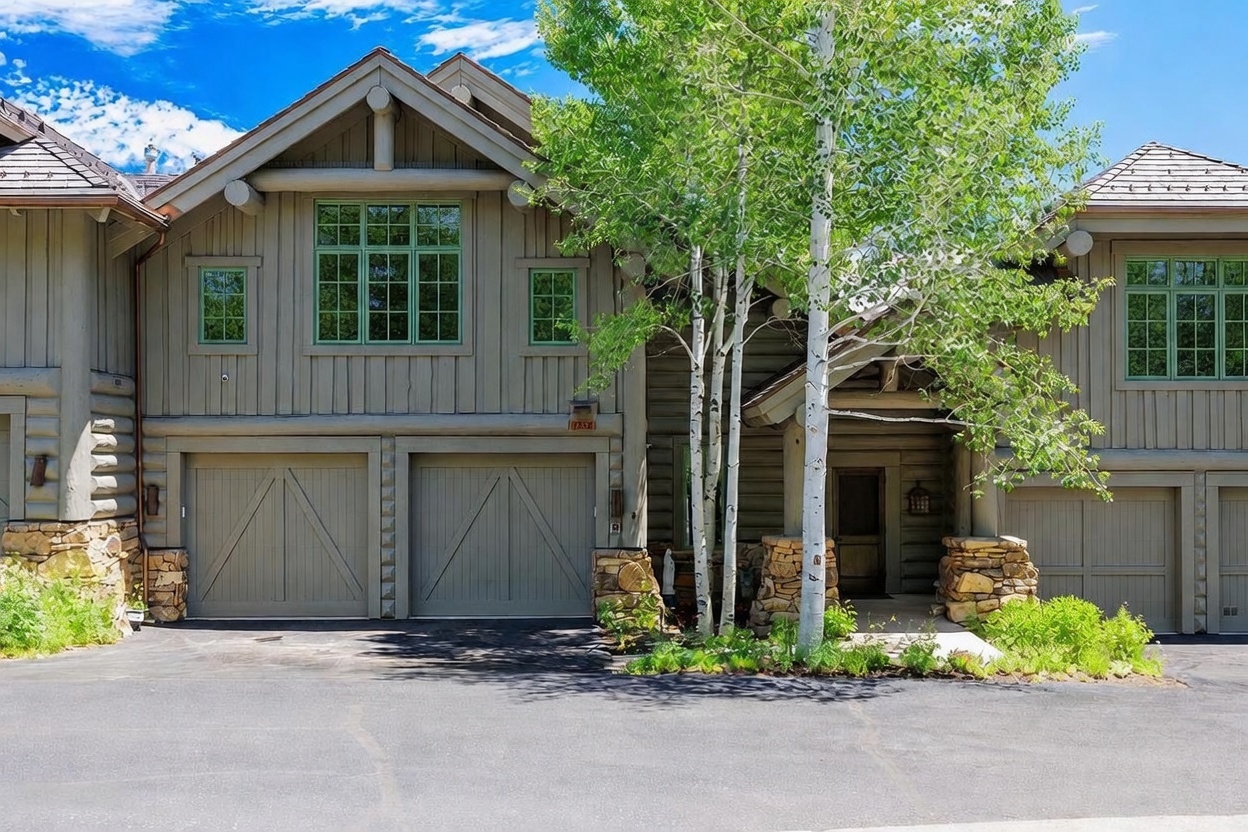 This front-facing view captures a multi-level mountain-style residence featuring a combination of vertical wood siding and rustic stone accents. The home includes a two-car garage with carriage-style doors, a covered entryway, and a prominent gable roofline. A cluster of slender birch trees stands in the foreground, adding a natural, textured element to the property's exterior facade.